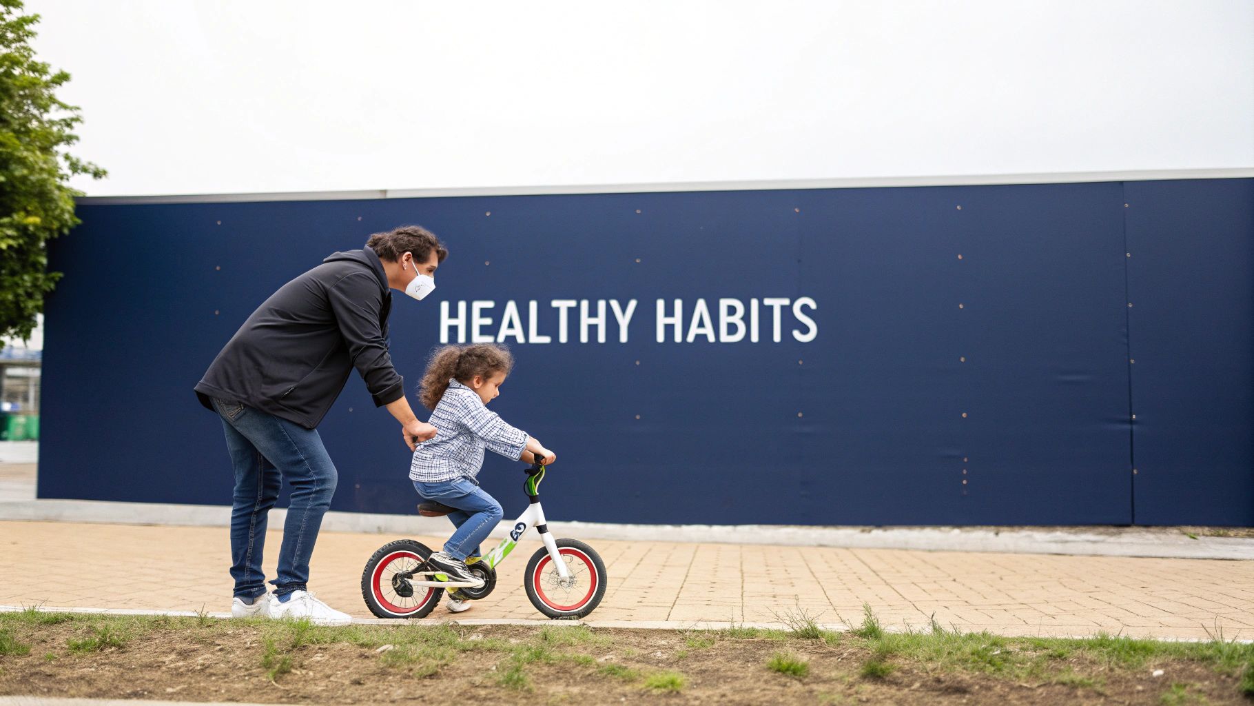 A man in a mask helps a child learn to ride a balance bike next to a 'HEALTHY HABITS' sign.