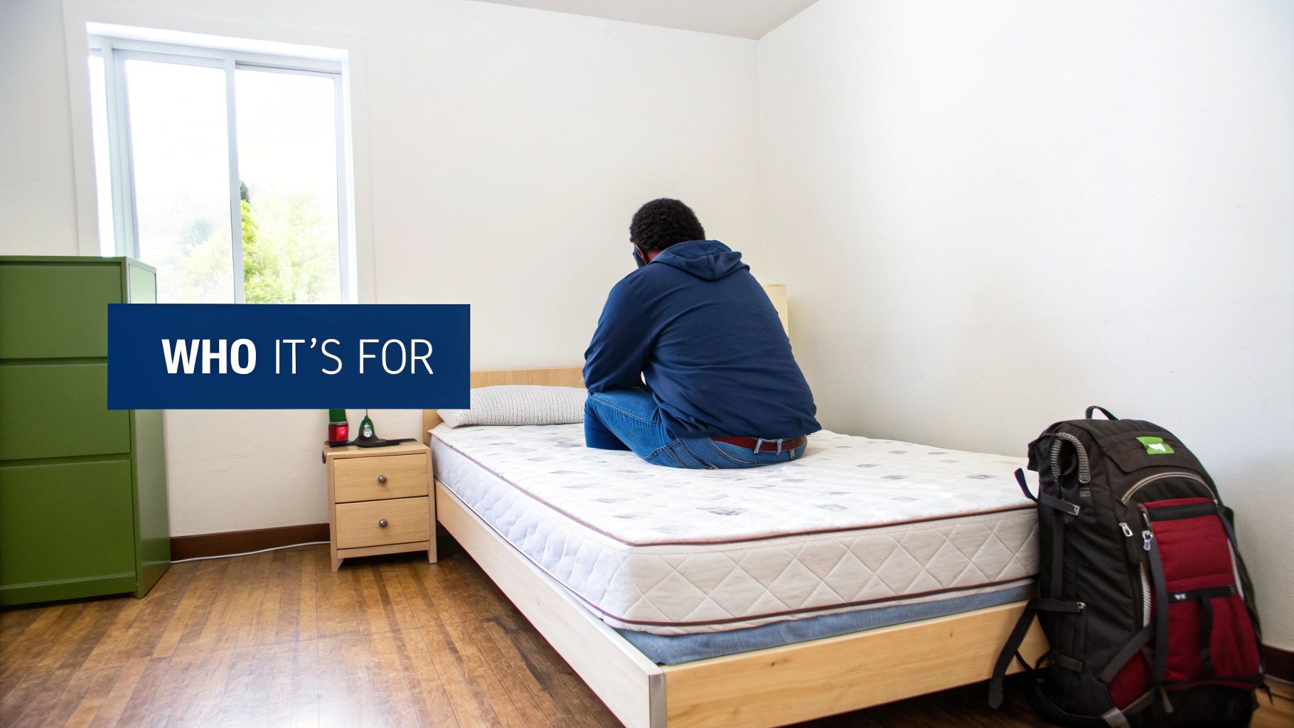Student sits on a new mattress in a simple dorm room, ready to move in.