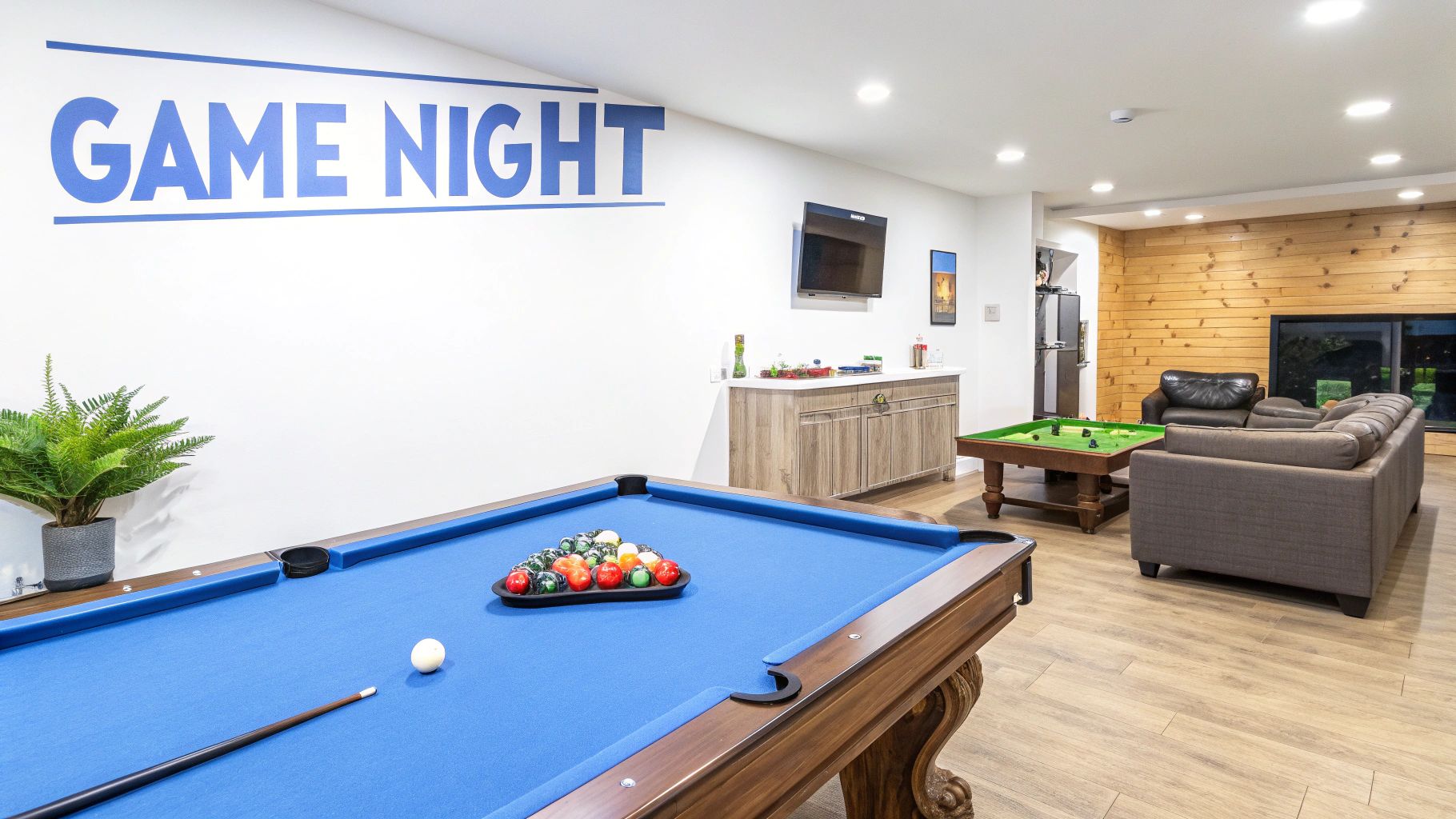 A picture of a finished basement game room with a pool table and wet bar, wood floors and a wall mounted TV.