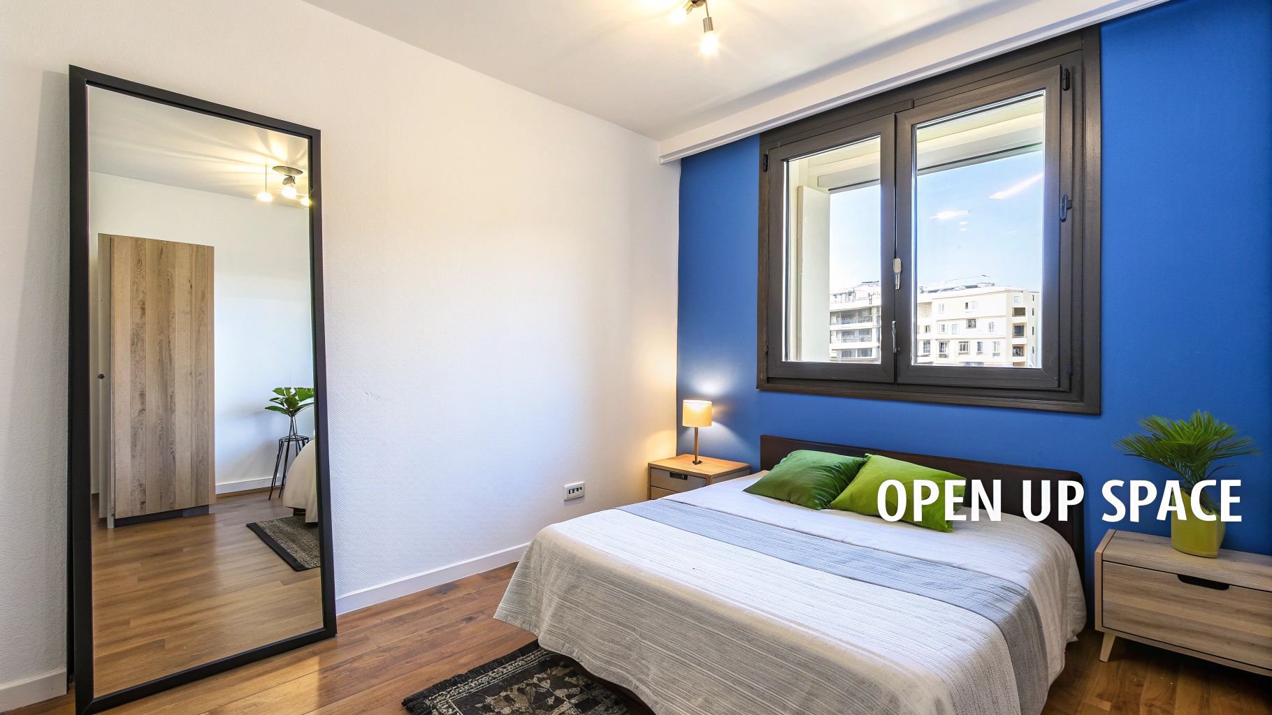 A modern, bright small bedroom featuring a bed with green pillows, a large mirror, and a blue accent wall.