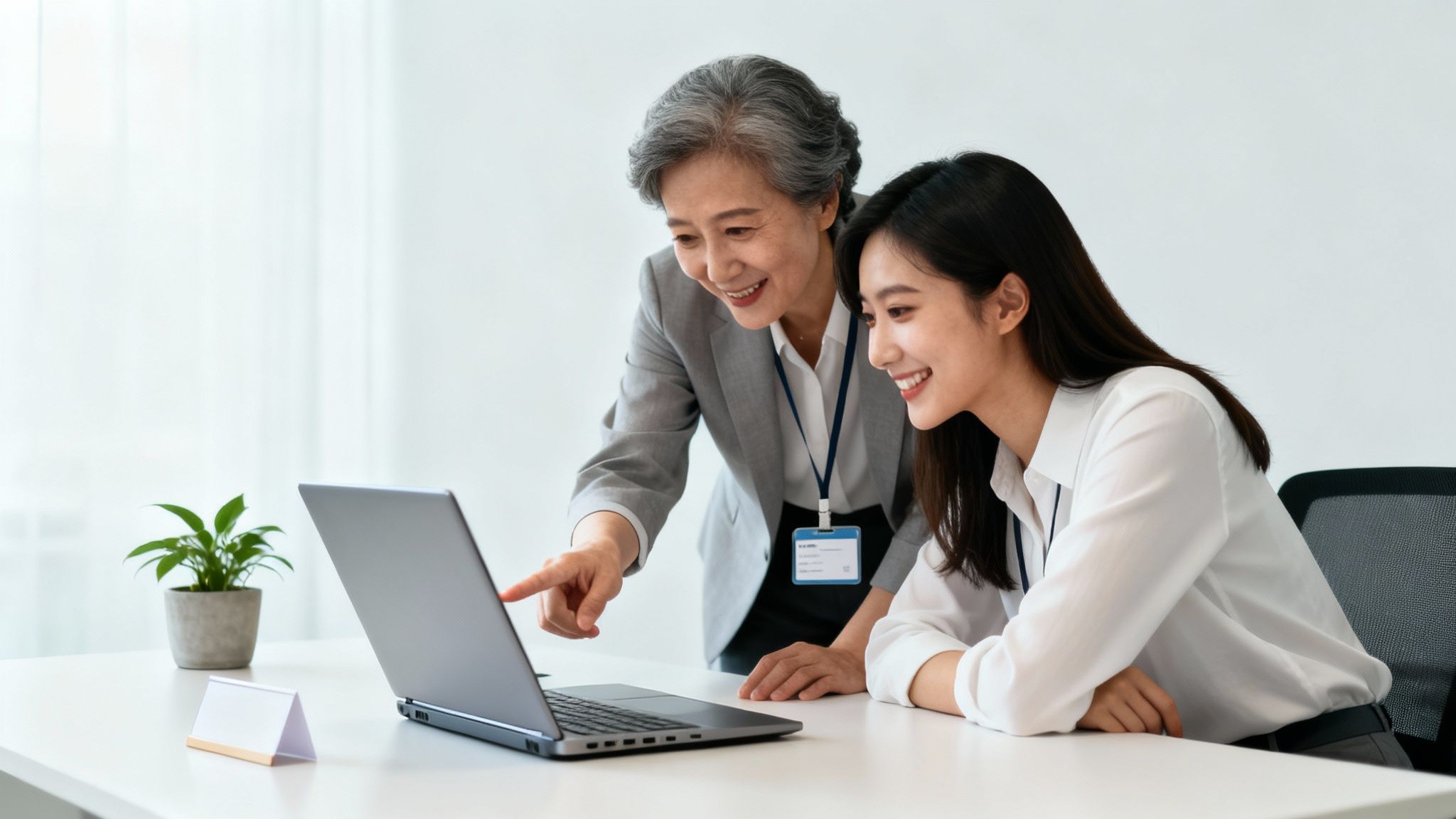 An older woman mentors a younger colleague, pointing at a laptop screen in a bright office.