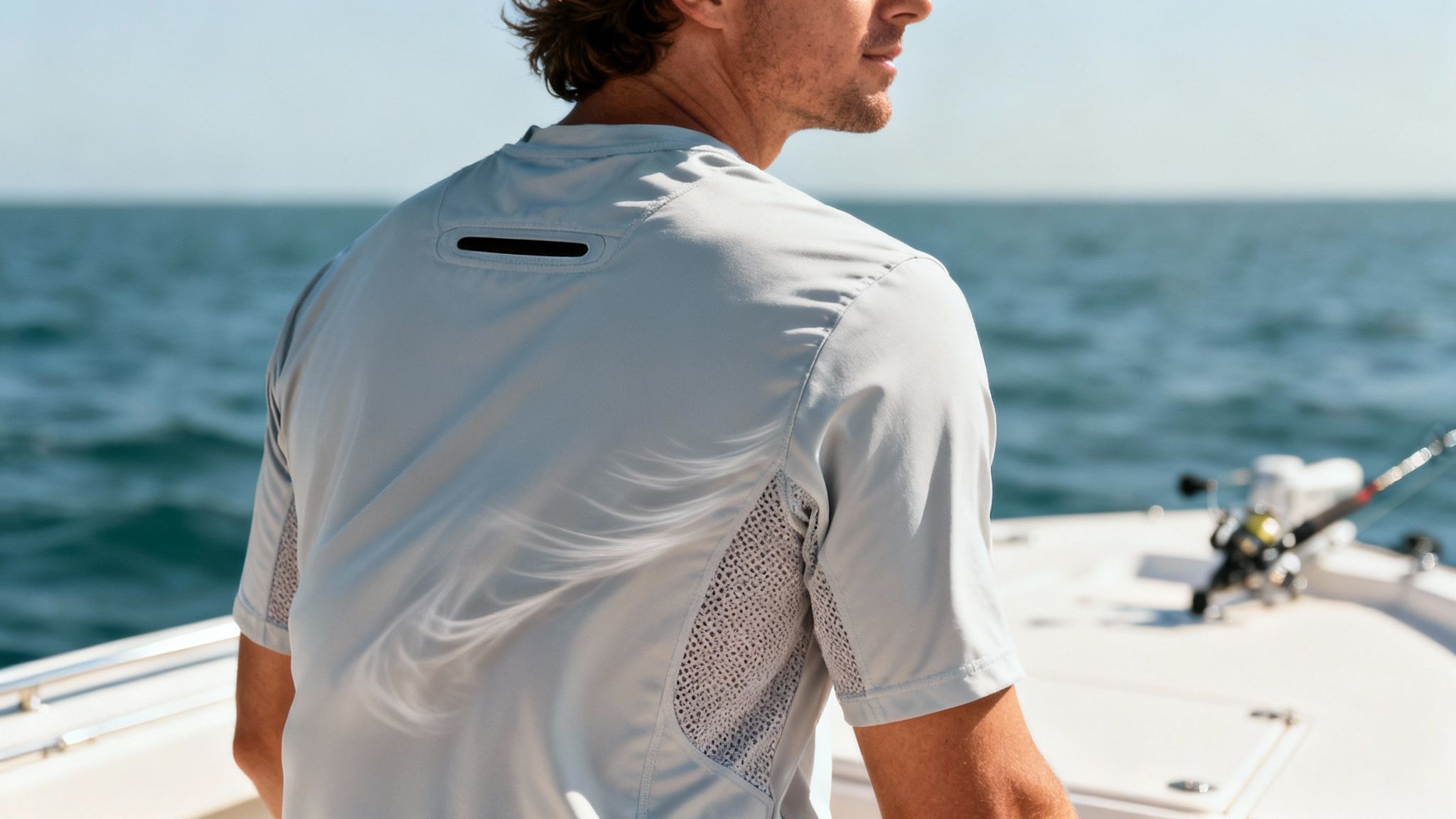 Back of a man in a light grey short-sleeve fishing shirt with mesh panels, looking at the ocean from a boat.