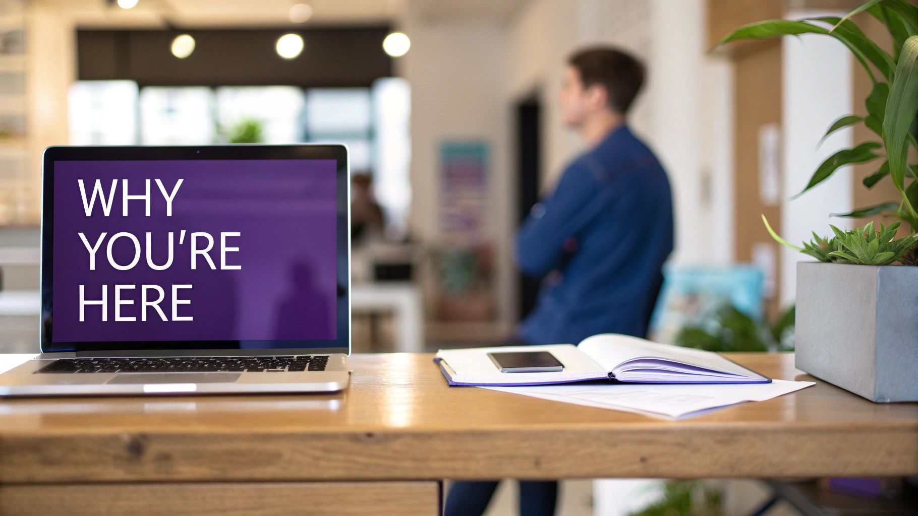 A laptop displays 'WHY YOU'RE HERE' on a desk with a notebook, phone, and plant in an office.