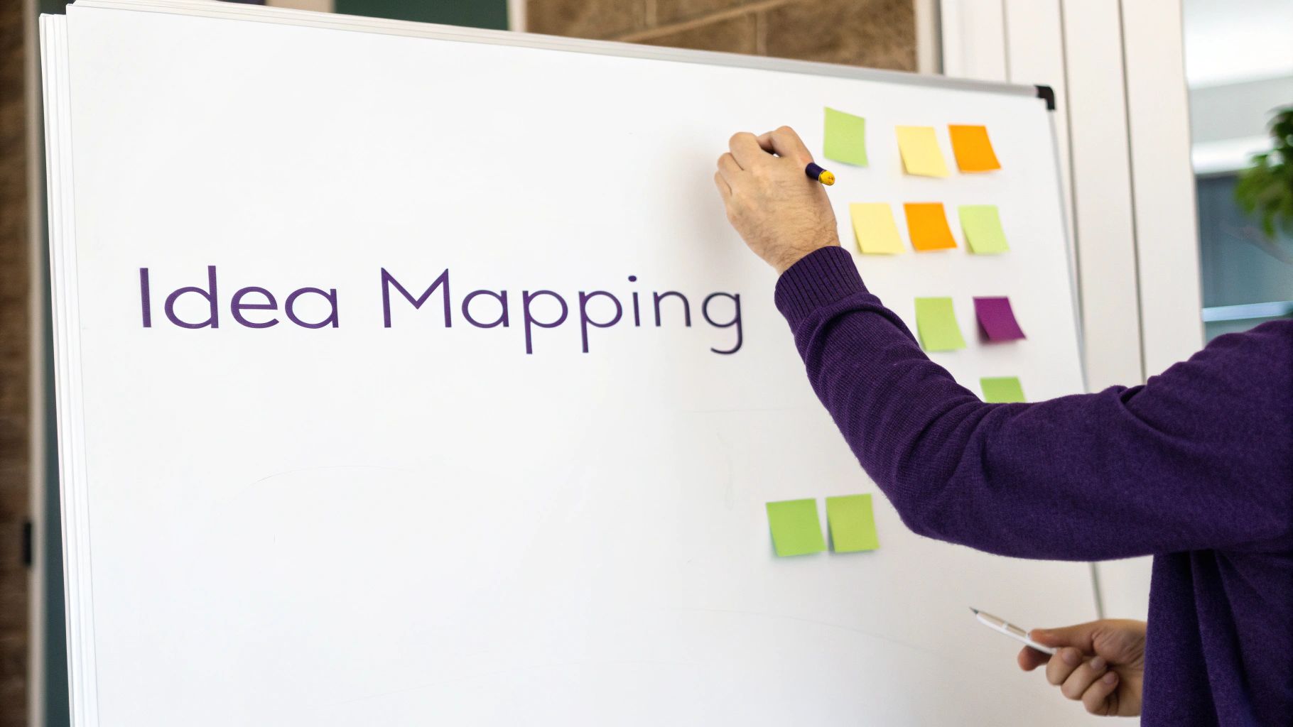 A person's hand writes 'Idea Mapping' on a whiteboard filled with colorful sticky notes.