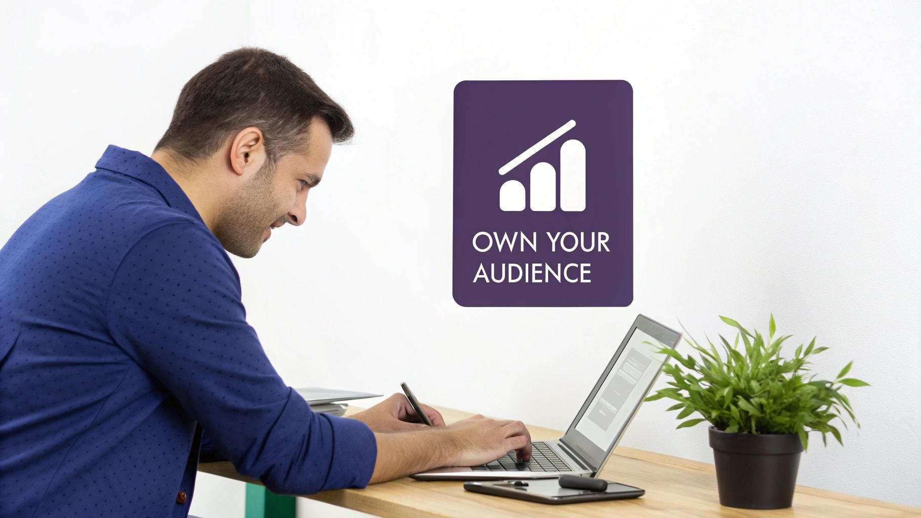 A man works on a laptop at a desk, with a sign "OWN YOUR AUDIENCE" in the background.