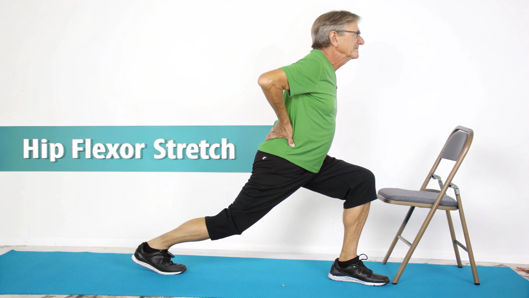 Senior man performing a hip flexor stretch on a blue yoga mat with a chair nearby.