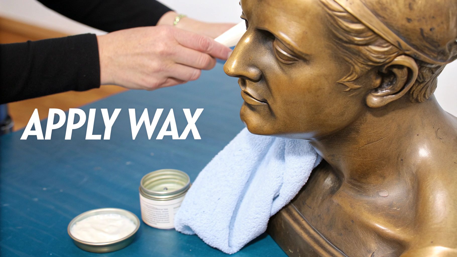 Hands carefully apply wax to a bronze bust, with jars of wax and a towel nearby.