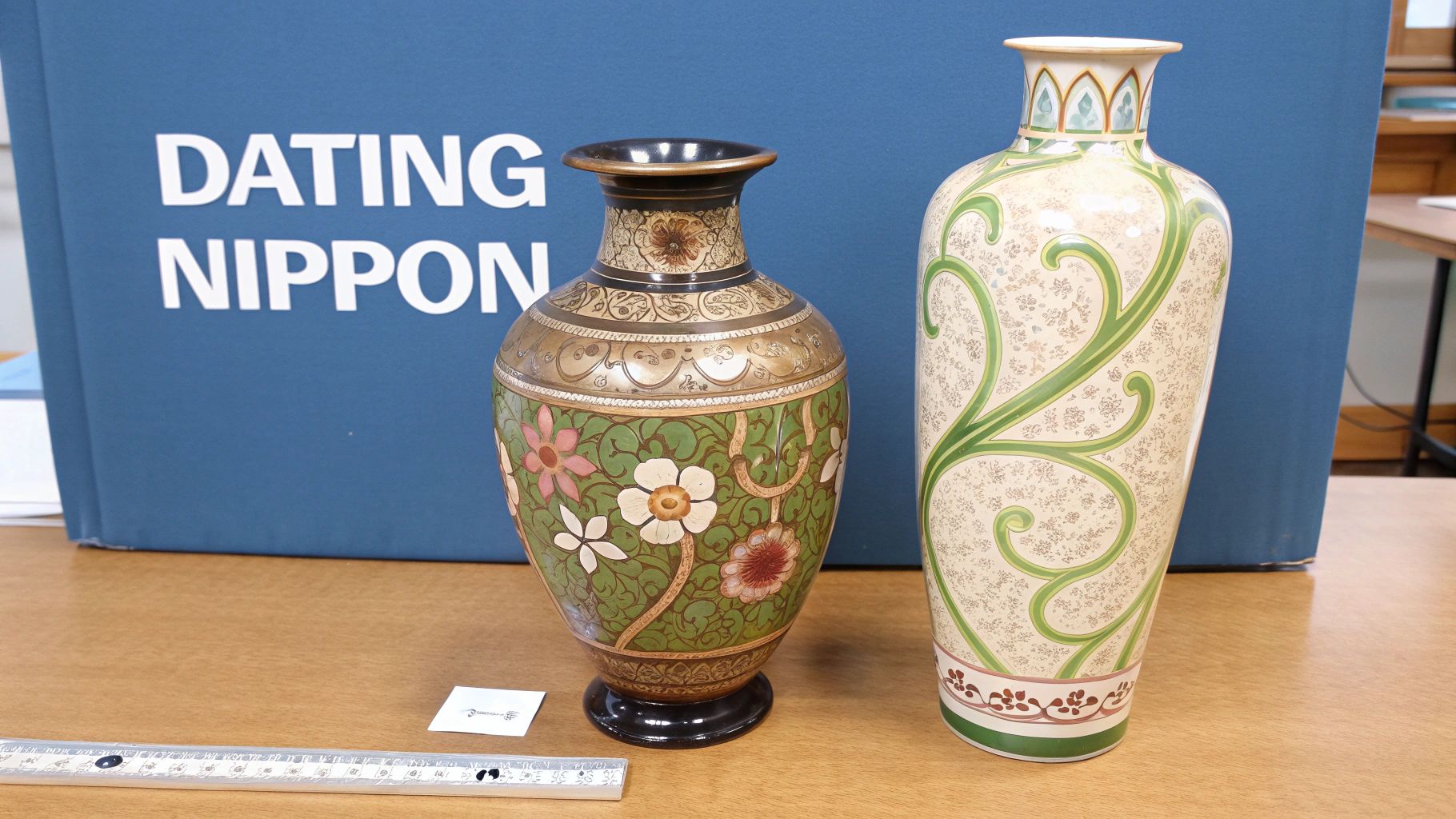 Two antique Japanese decorative vases with intricate floral and scroll patterns on a table with a ruler.