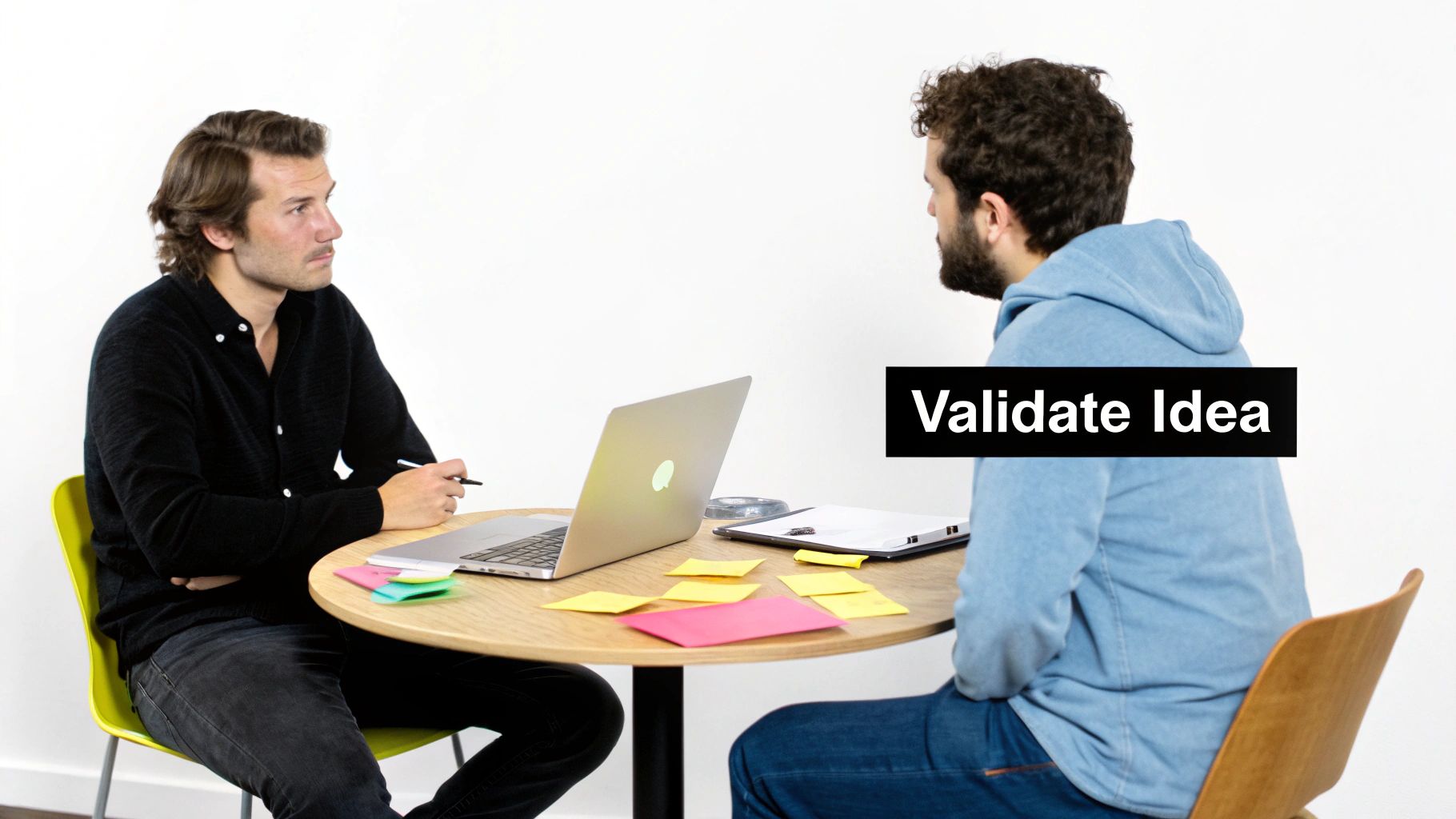 Two men discuss a project, with a laptop and sticky notes on a table, validating an idea.