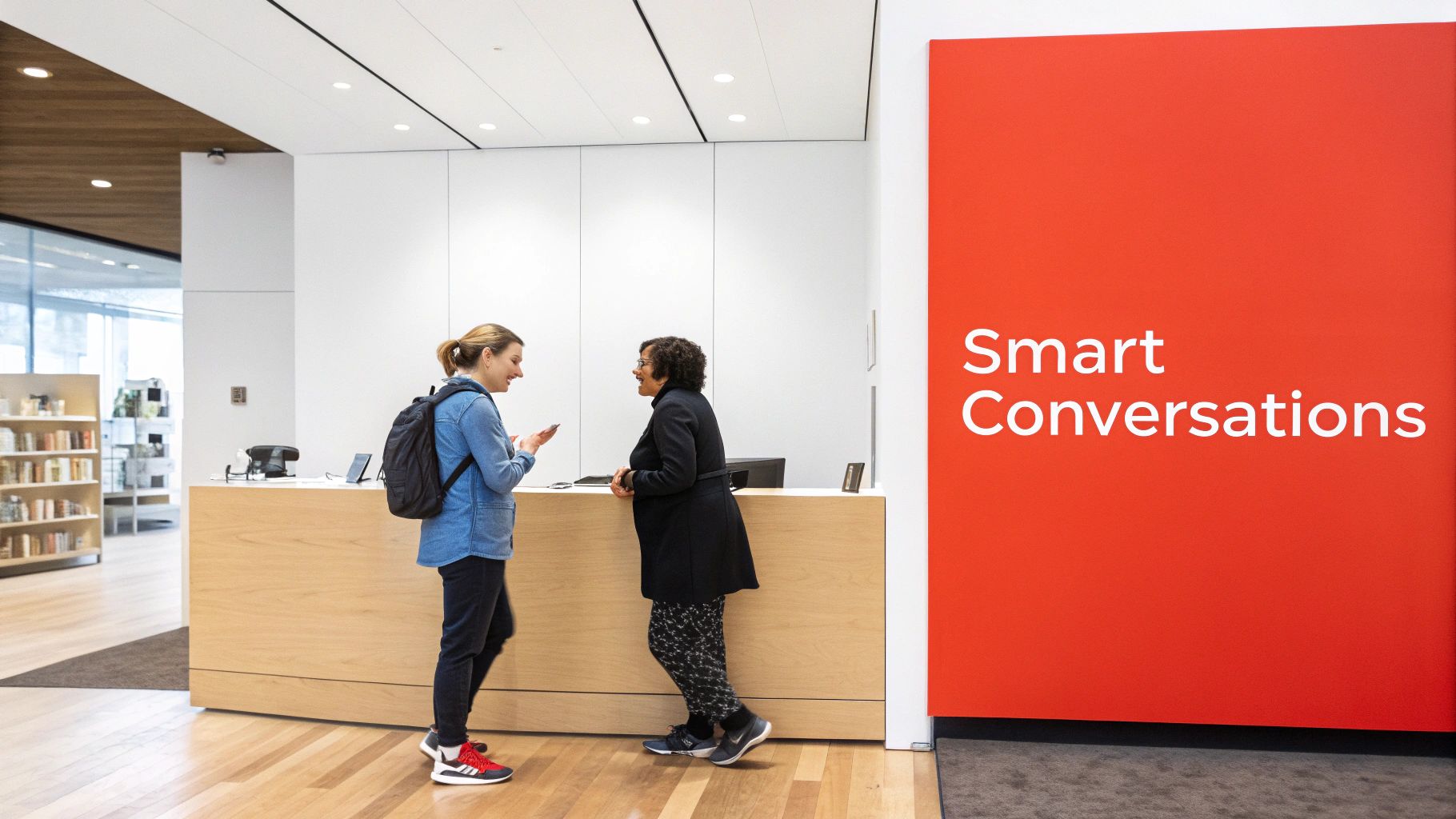 Two women chat at a wooden reception desk in a modern office with a 'Smart Conversations' sign.