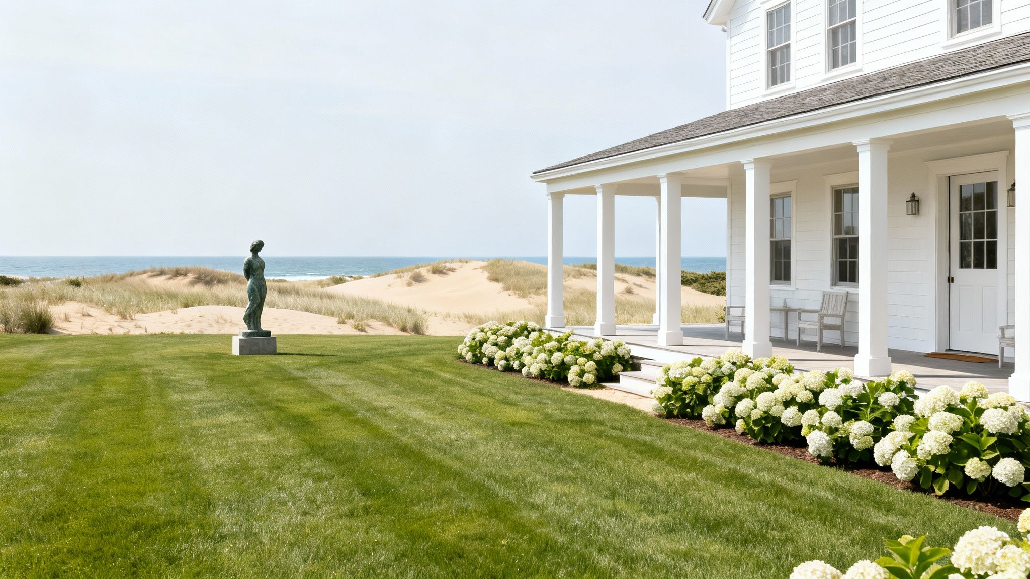 A luxurious white Hamptons home with a grand porch, green lawn, and a bronze statue overlooking sand dunes and the ocean.