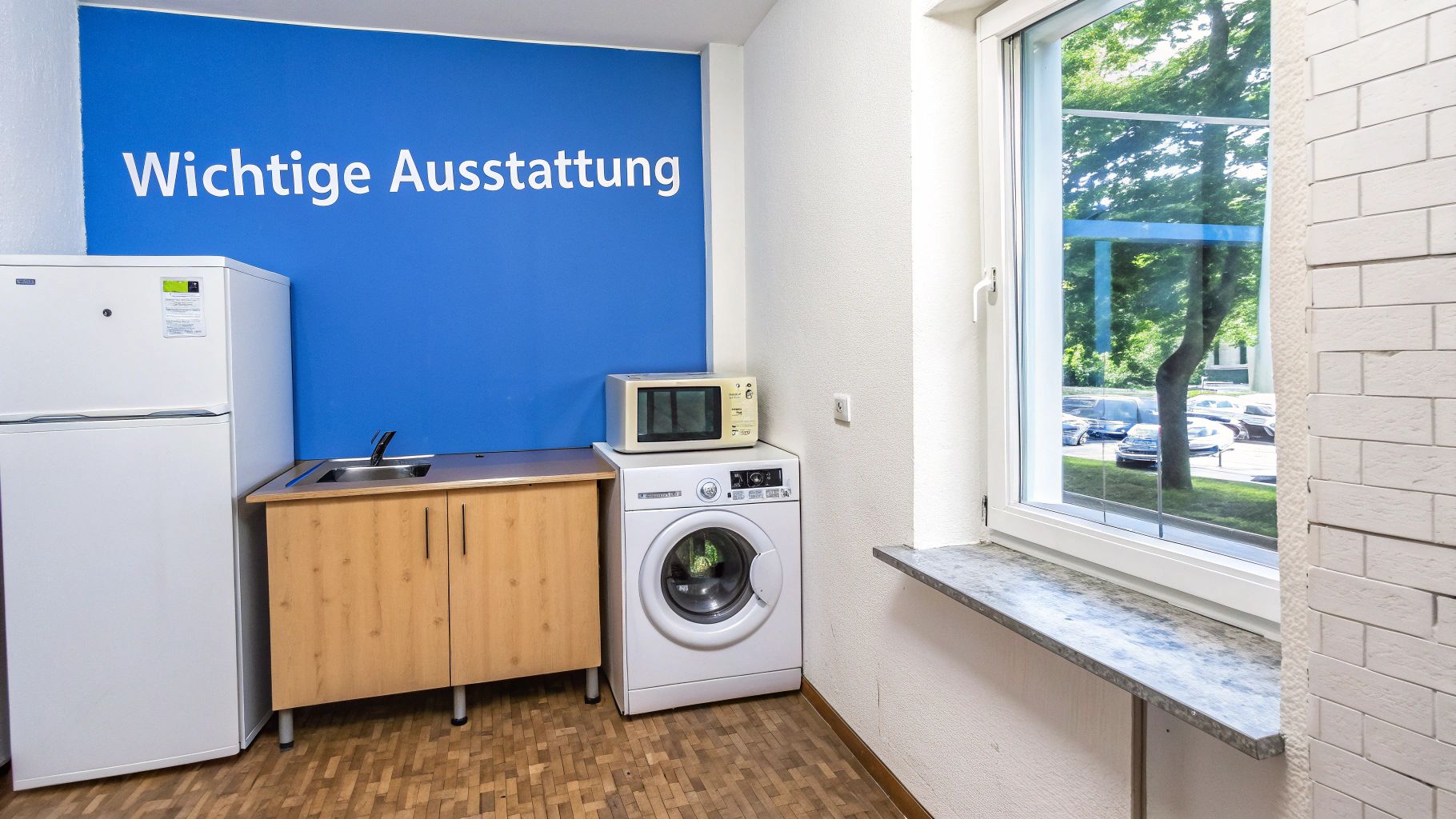 Kleine Küchenzeile mit Kühlschrank, Spüle, Schränken, Waschmaschine und Mikrowelle vor einer blauen Wand mit Text. Ein Fenster bietet Blick nach draußen.