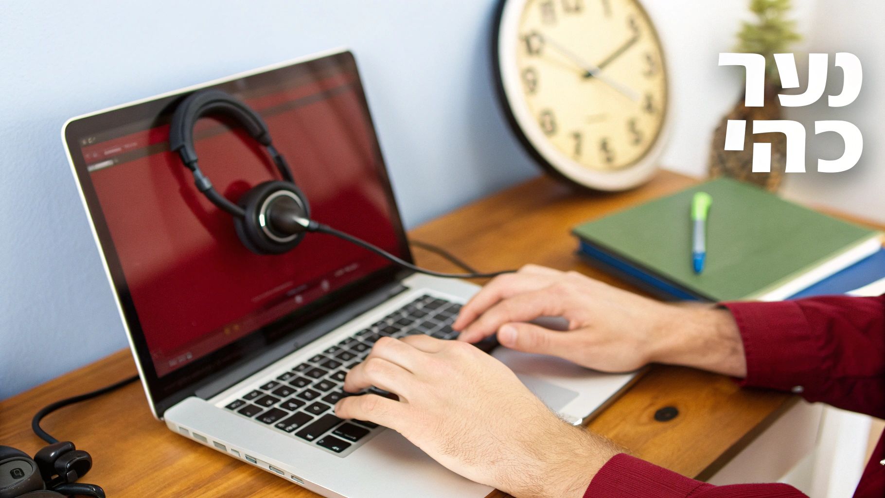 אדם מקליד במחשב נייד עם אוזניות על המסך, יושב ליד שעון וספרים על שולחן עץ.