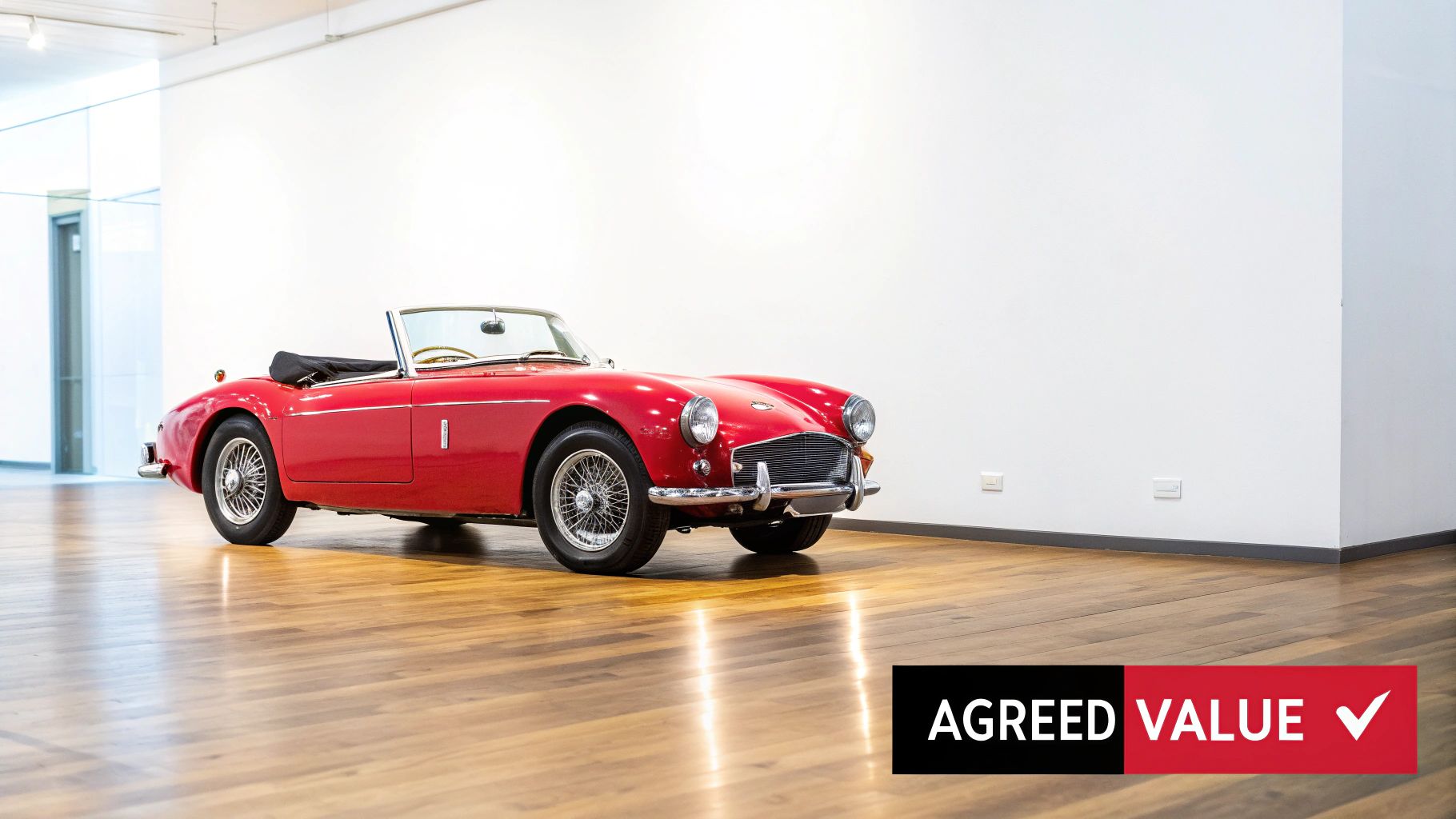 A gleaming red vintage convertible sports car is parked on a polished wooden floor in a minimalist room.