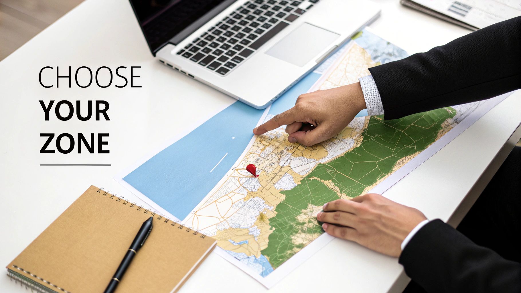 A person's hands pointing at a map with a red pin, next to a laptop and a notebook with text 'CHOOSE YOUR ZONE'.