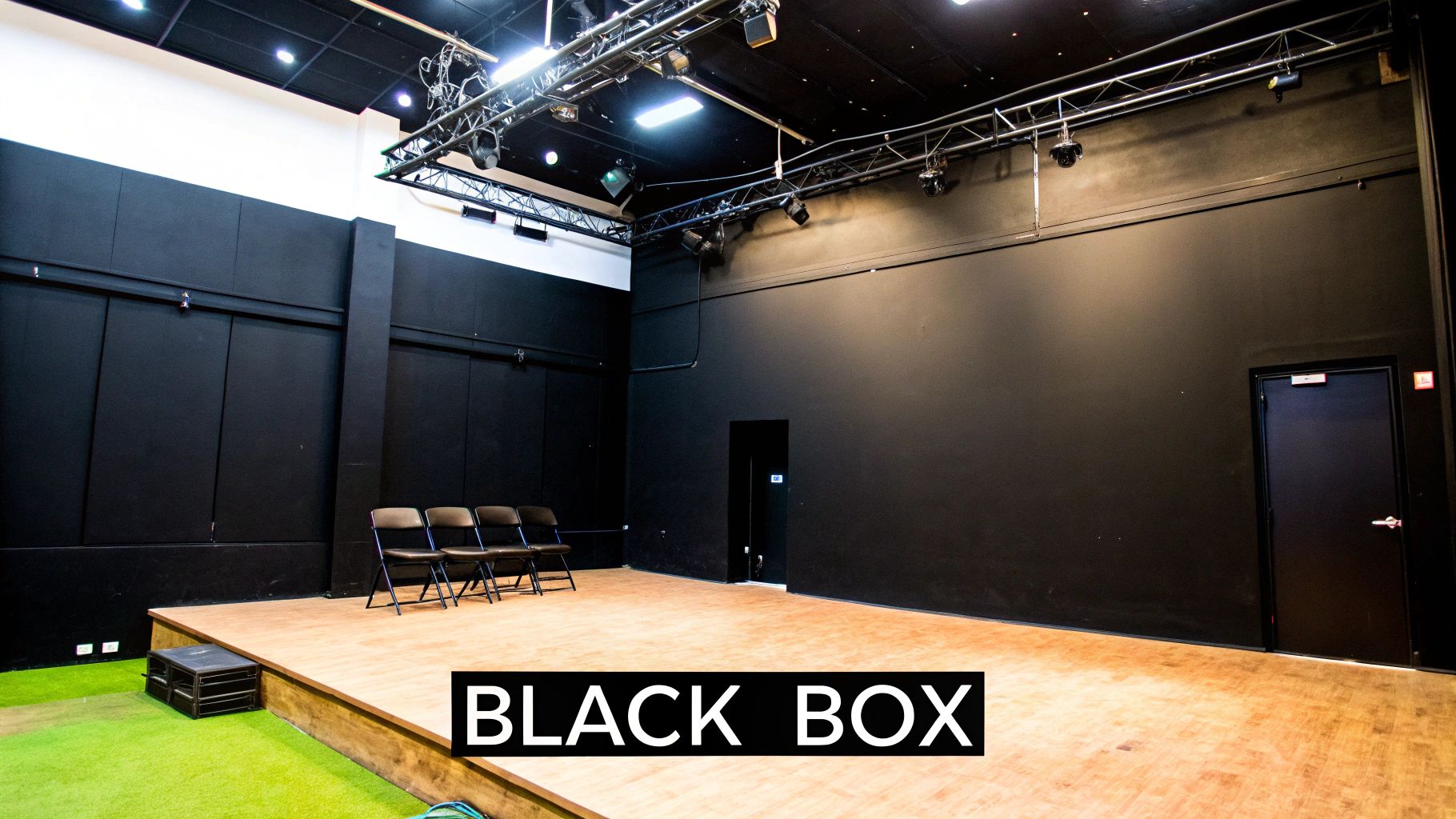 An empty black box theater with a wooden stage, overhead lighting trusses, and black walls.