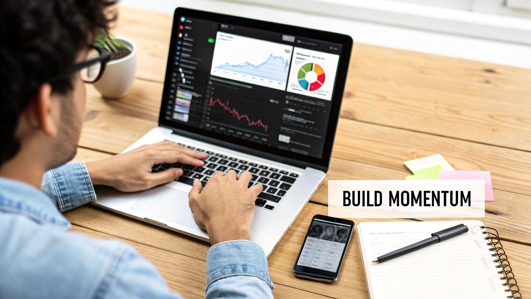 Man working on a laptop with financial charts and a smartphone on a wooden desk with a notebook.