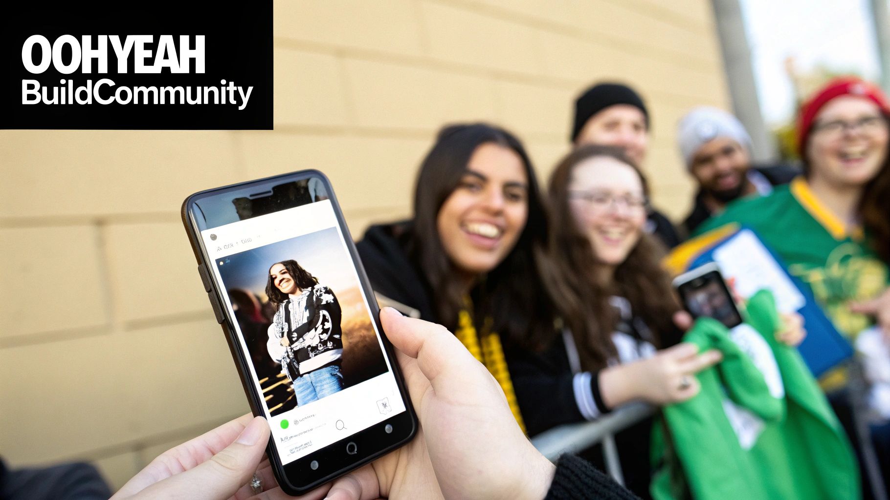 A person holds a smartphone displaying a photo of a musician, with a diverse crowd of smiling fans in the background.