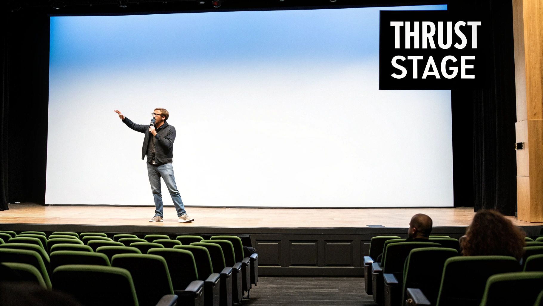 A man speaks into a microphone on a thrust stage with a large screen and green auditorium seats.