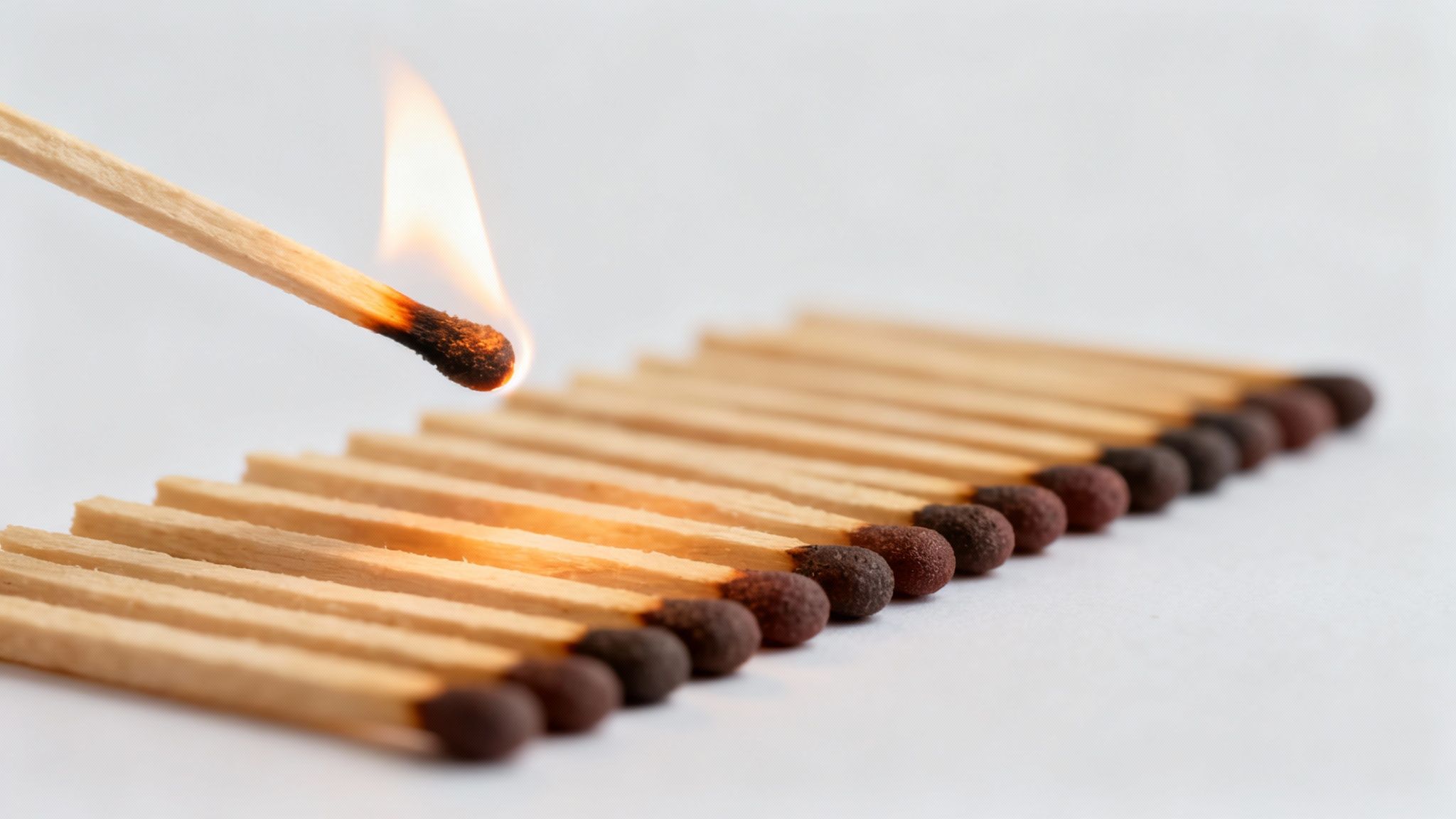 A lit matchstick igniting the first of a row of unlit matches on a white background.