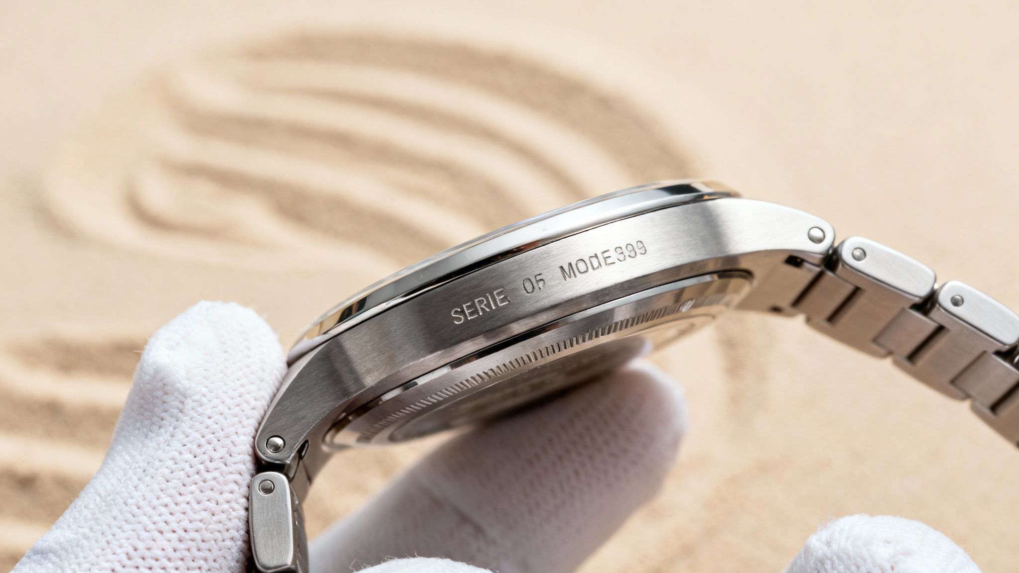 A gloved hand holds a silver watch with 'SERIE 05 MODE399' engraved on its side, against sand.