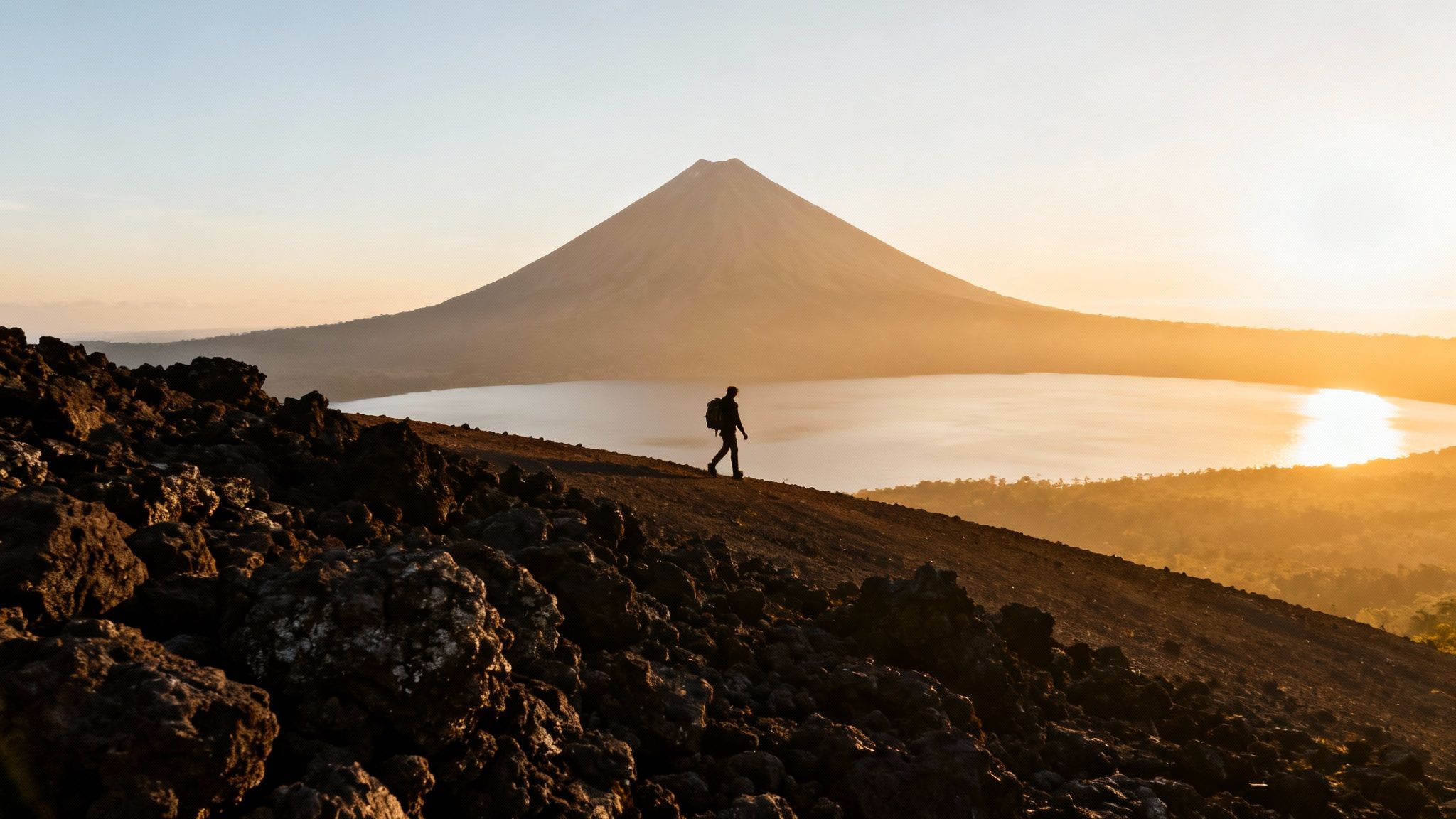 Best hikes in Costa Rica — silhouetted hiker on a rocky mountain trail with a volcano and sunlit lake in the background.