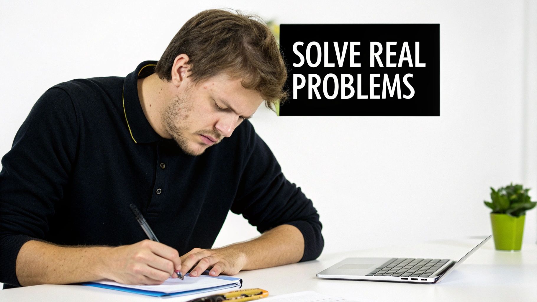 A focused man in a black polo shirt writes notes at a modern white desk.