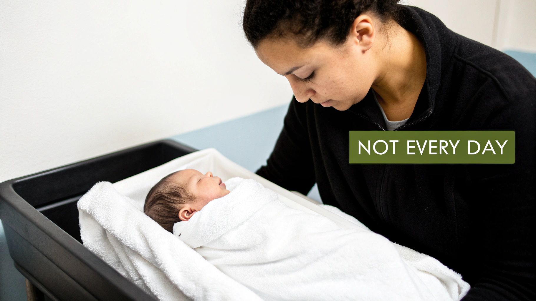 A woman gently looks down at her newborn baby, wrapped in a white towel in a dark basin.