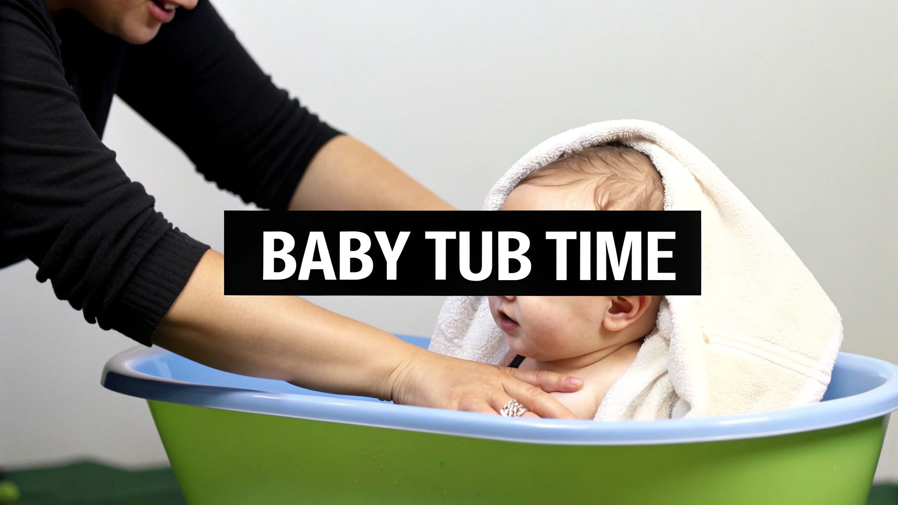 An adult's hands gently assist a baby wrapped in a soft white towel in a green and blue bath tub.