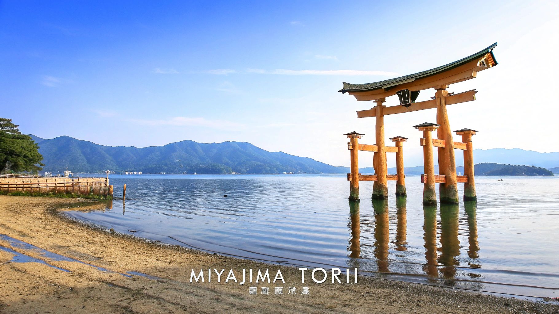Iconic orange Miyajima Torii gate standing in calm blue water with distant mountains and a beach.