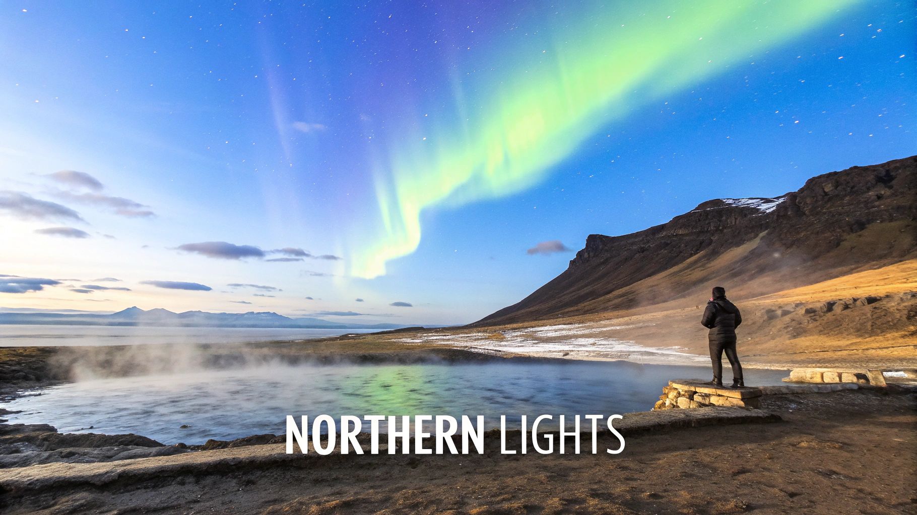 A lone traveler watches the vibrant green Northern Lights illuminate a steaming lake and majestic mountains.
