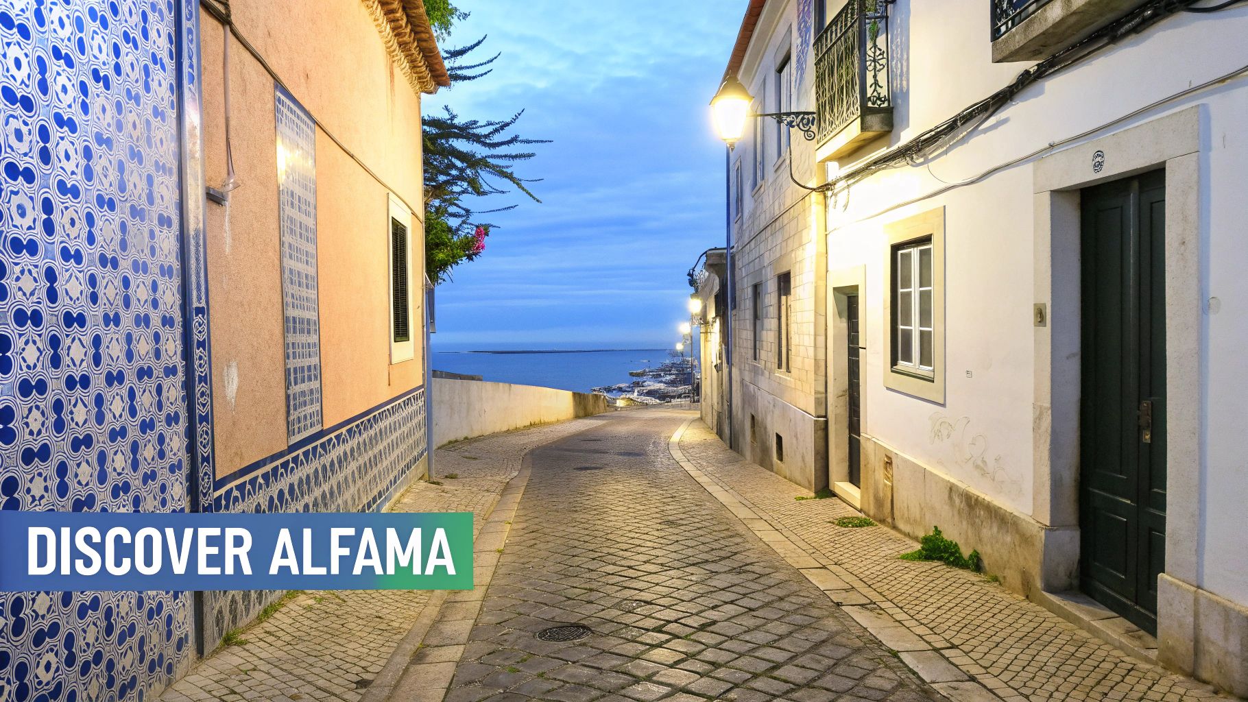 A charming, cobbled street in Alfama, Lisbon, illuminated by streetlights at dusk, leading to the Tagus River.