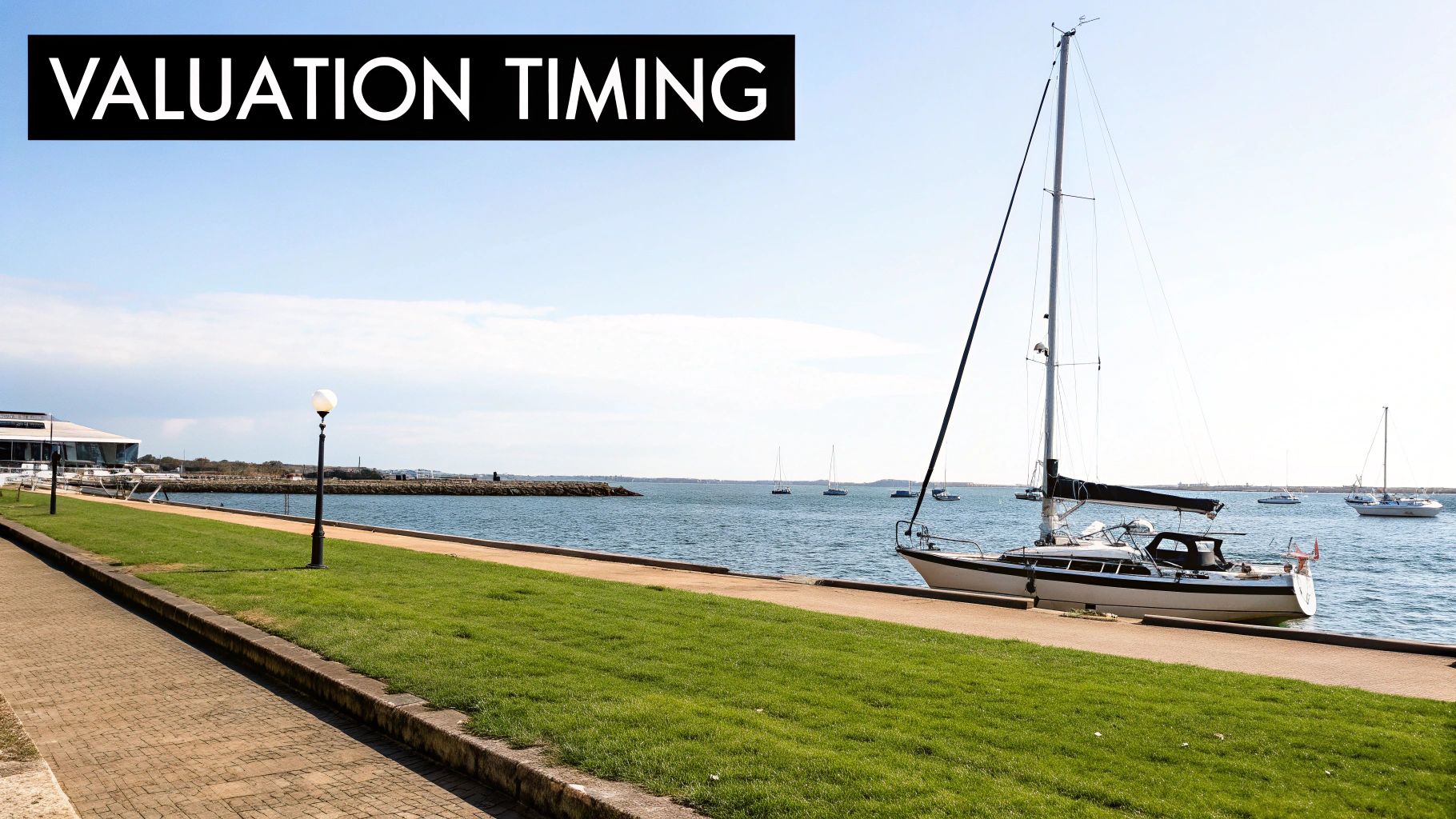 A scenic waterfront with a paved path, green grass, a docked sailboat, and boats on the water.