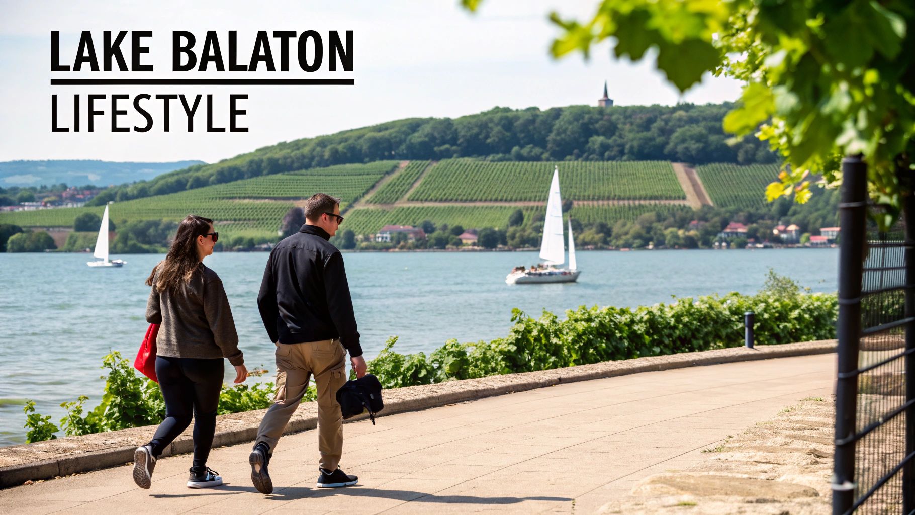 A couple walks along a sunny promenade by Lake Balaton, with sailboats and vineyards in the background.