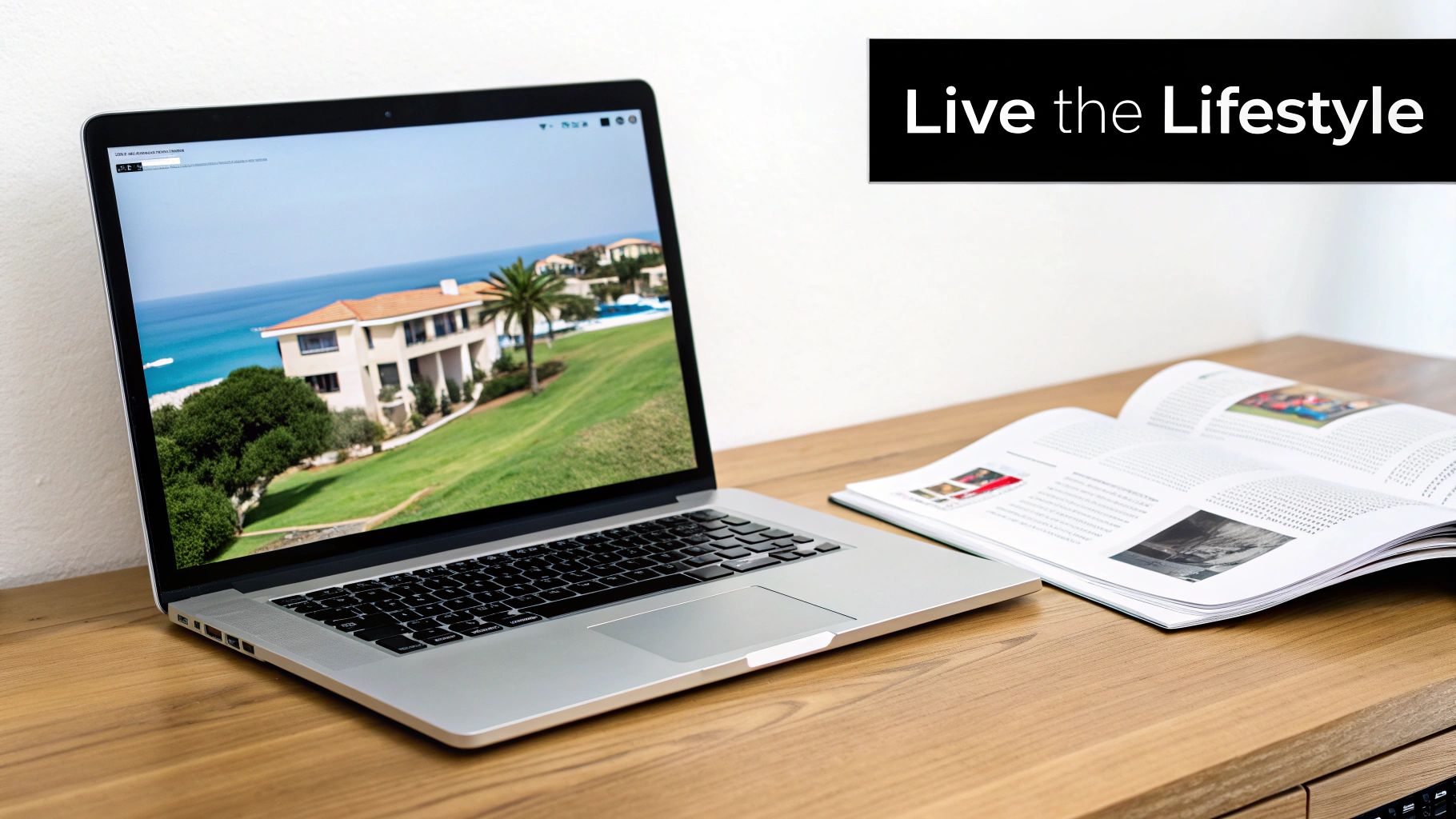 A laptop displays a luxury villa with a sea view, beside an open lifestyle magazine on a wooden desk.