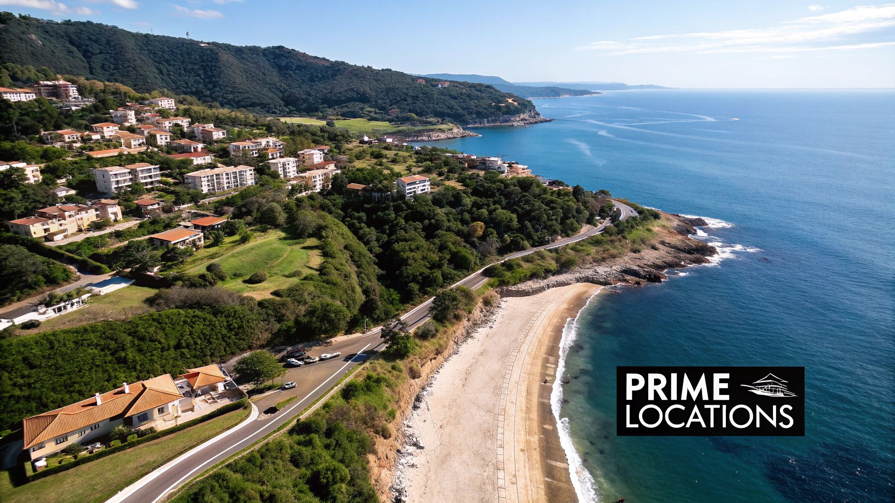Aerial view of a scenic coastline with hillside houses, a winding road, sandy beach, and clear blue sea.