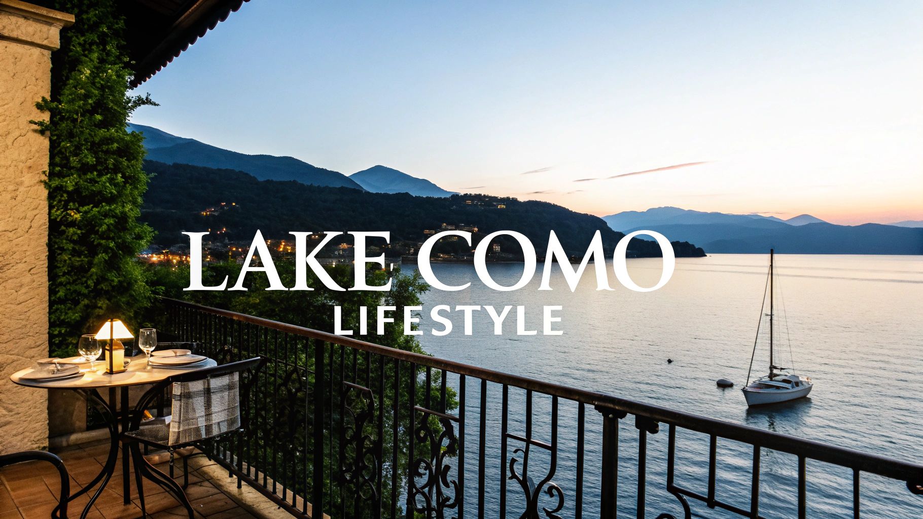 A romantic balcony view of Lake Como at sunset, with mountains, a sailboat, and a table set for two.