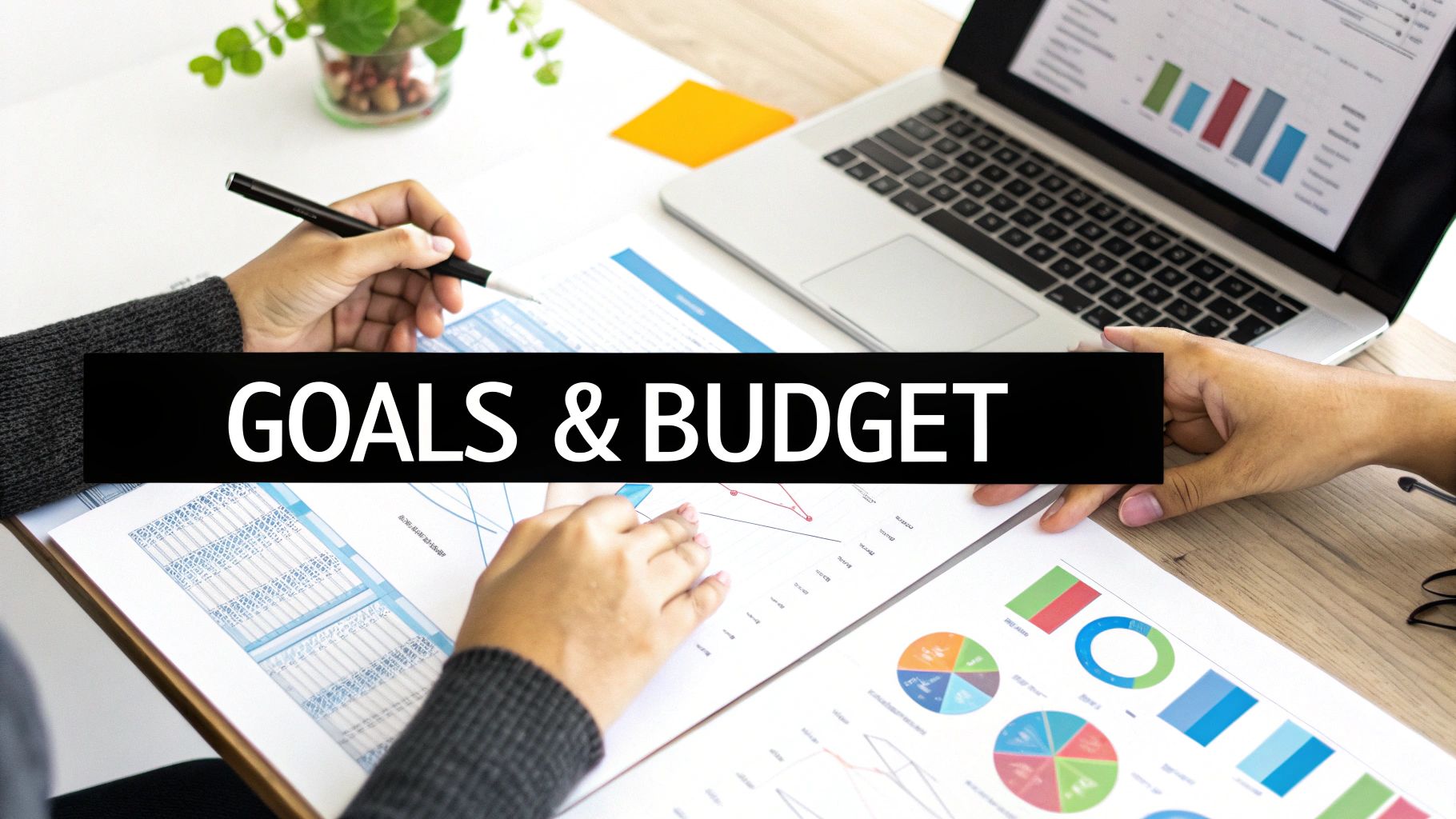 A person writing down goals and budget information in a notebook, with a laptop and coffee nearby.