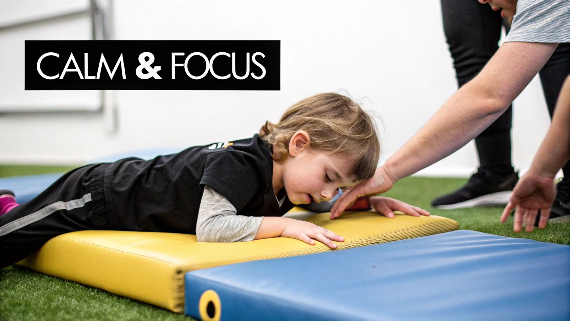 A young child lies focused on a yellow sensory mat with an adult's hand gently assisting.
