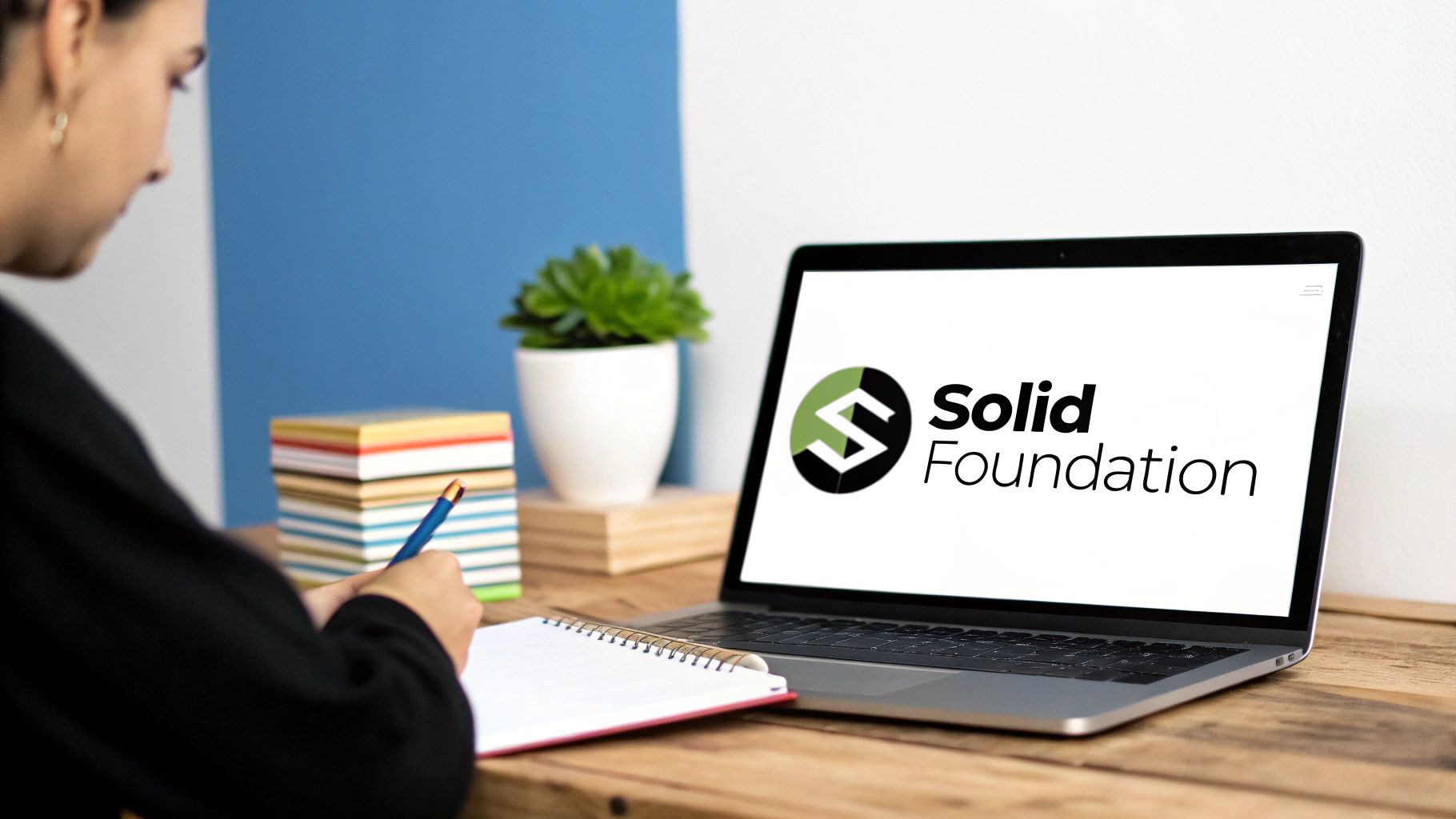 A person writes in a notebook at a wooden desk with a laptop showing the Solid Foundation logo.