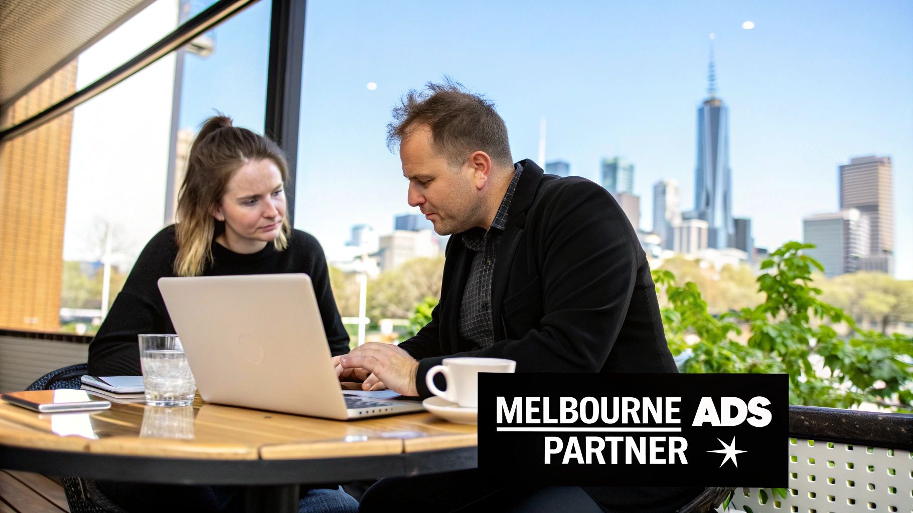 Two people, a man and a woman, working on a laptop at an outdoor table with the Melbourne skyline in the background.