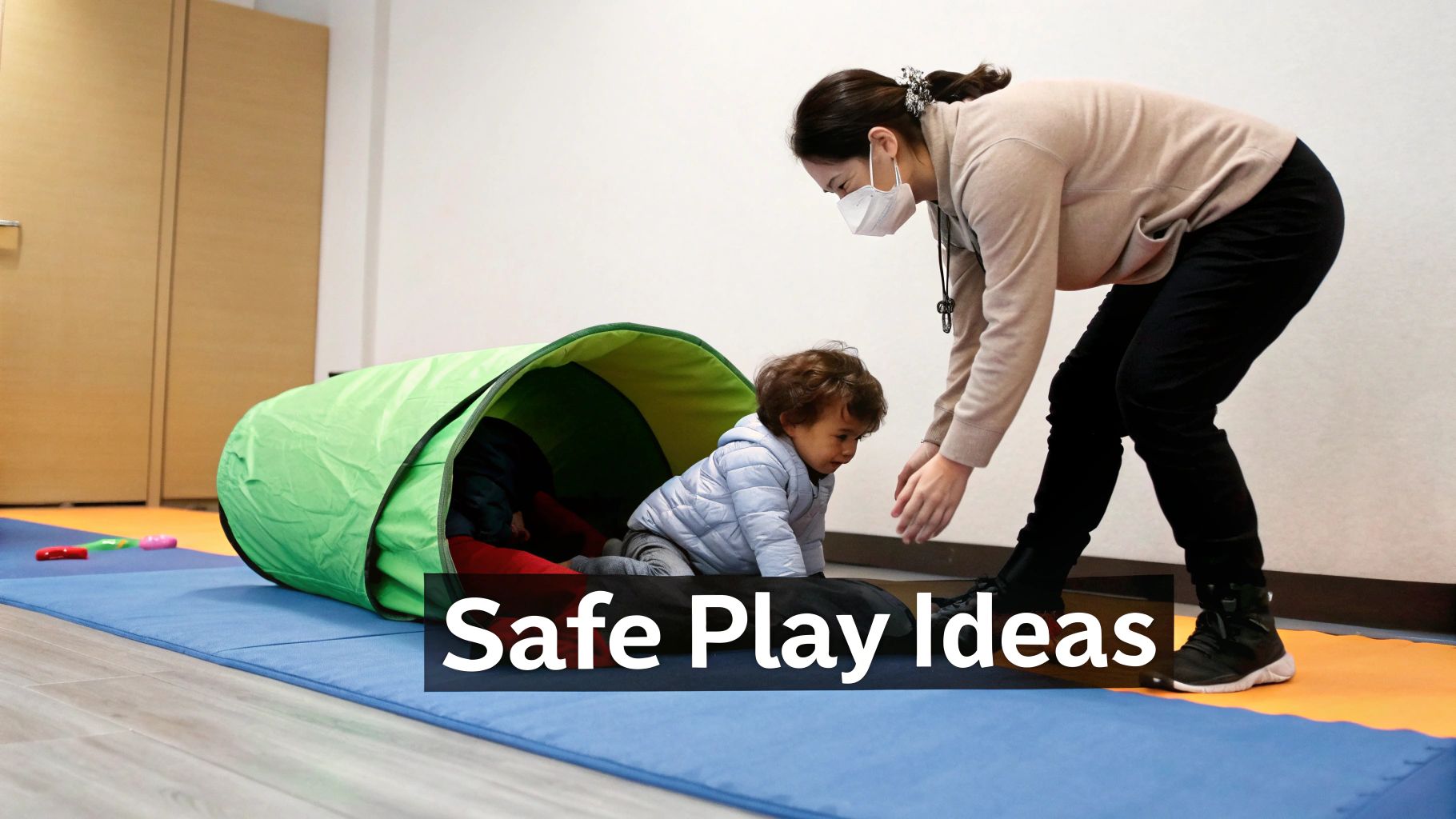 An adult wearing a mask helps a child crawl from a green play tunnel on soft mats.