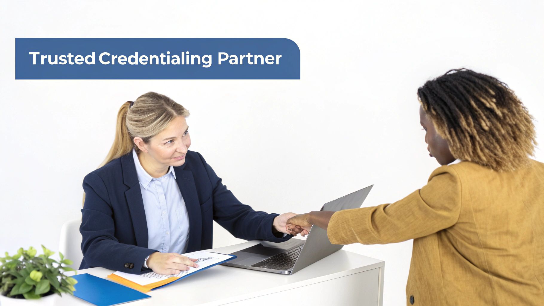 Two businesswomen shaking hands over a laptop and documents on a white desk, representing partnership.
