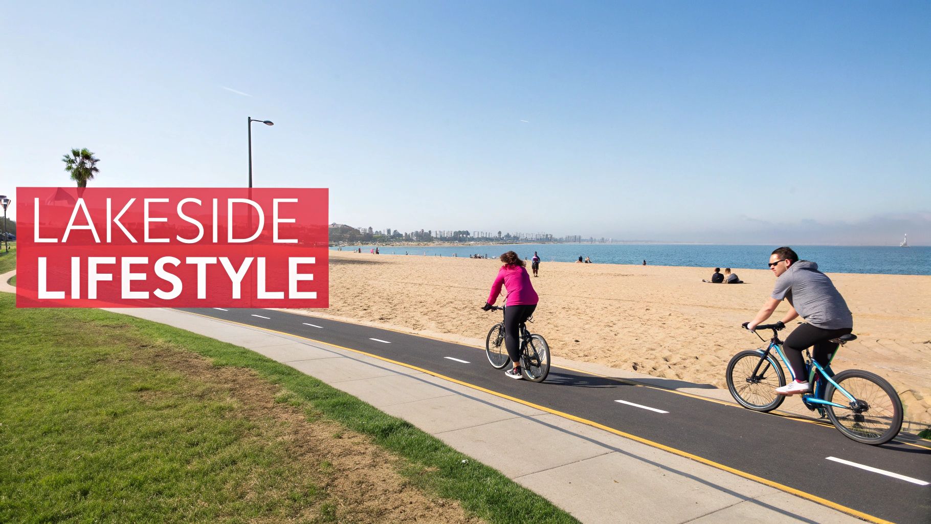 Two people cycling on a paved path next to a sandy beach and the ocean under a clear sky.