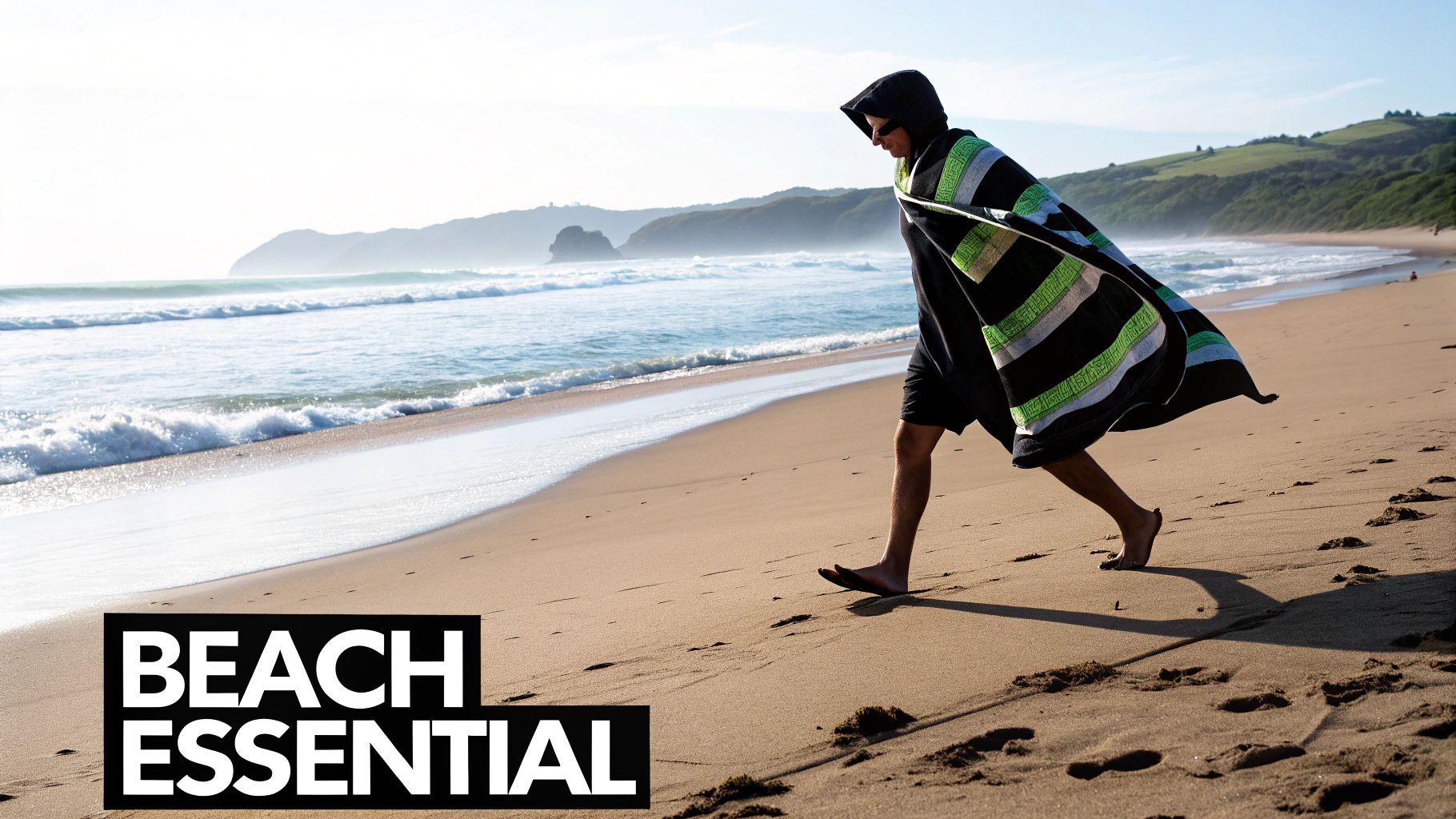 A person wearing a striped hooded beach poncho walks barefoot on a sandy beach next to ocean waves.