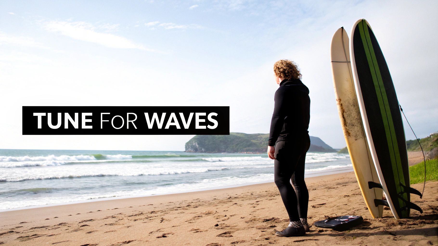 A surfer in a wetsuit stands on a sandy beach, looking at the waves with two surfboards beside them.