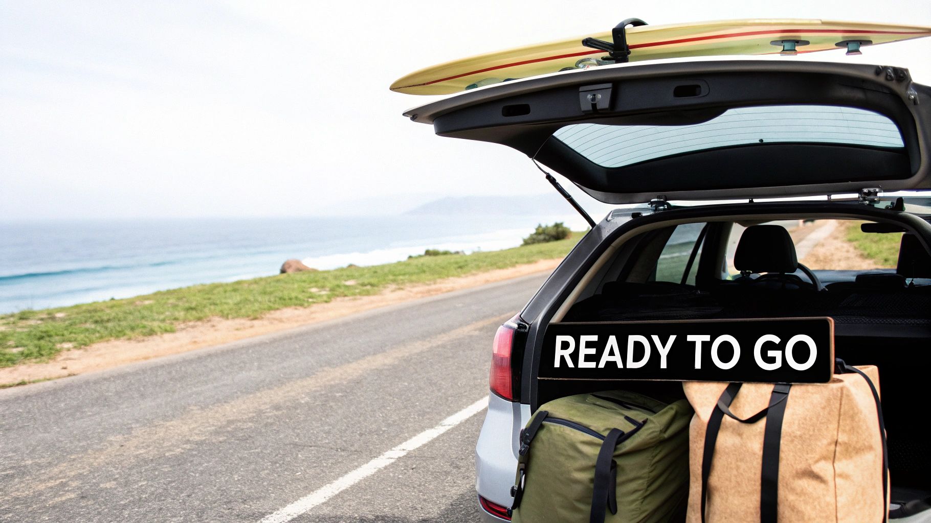 Car by the ocean with packed bags, a surfboard on the roof, and a "READY TO GO" sign in the trunk.