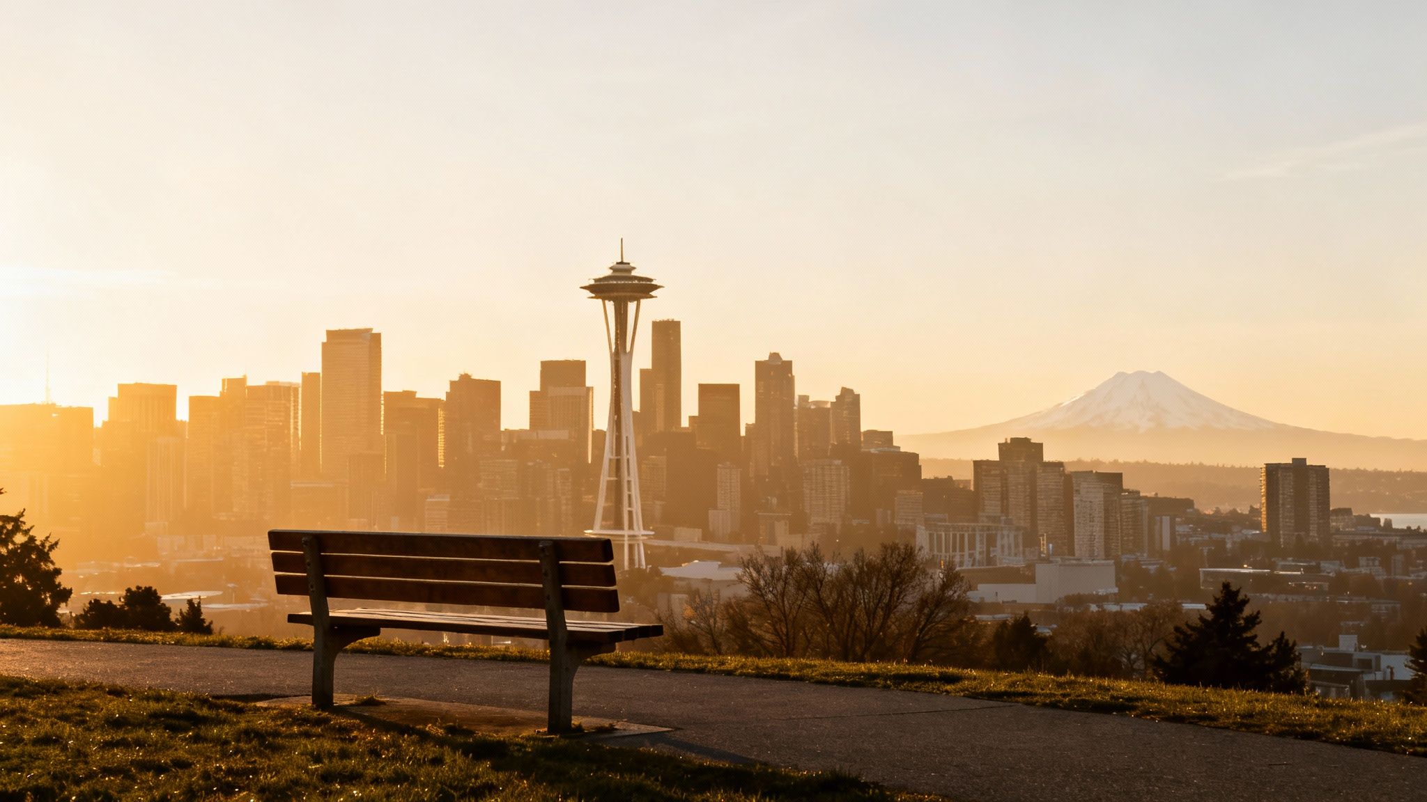 A picturesque golden hour view of the Seattle skyline, Space Needle, and Mount Rainier from a park bench.