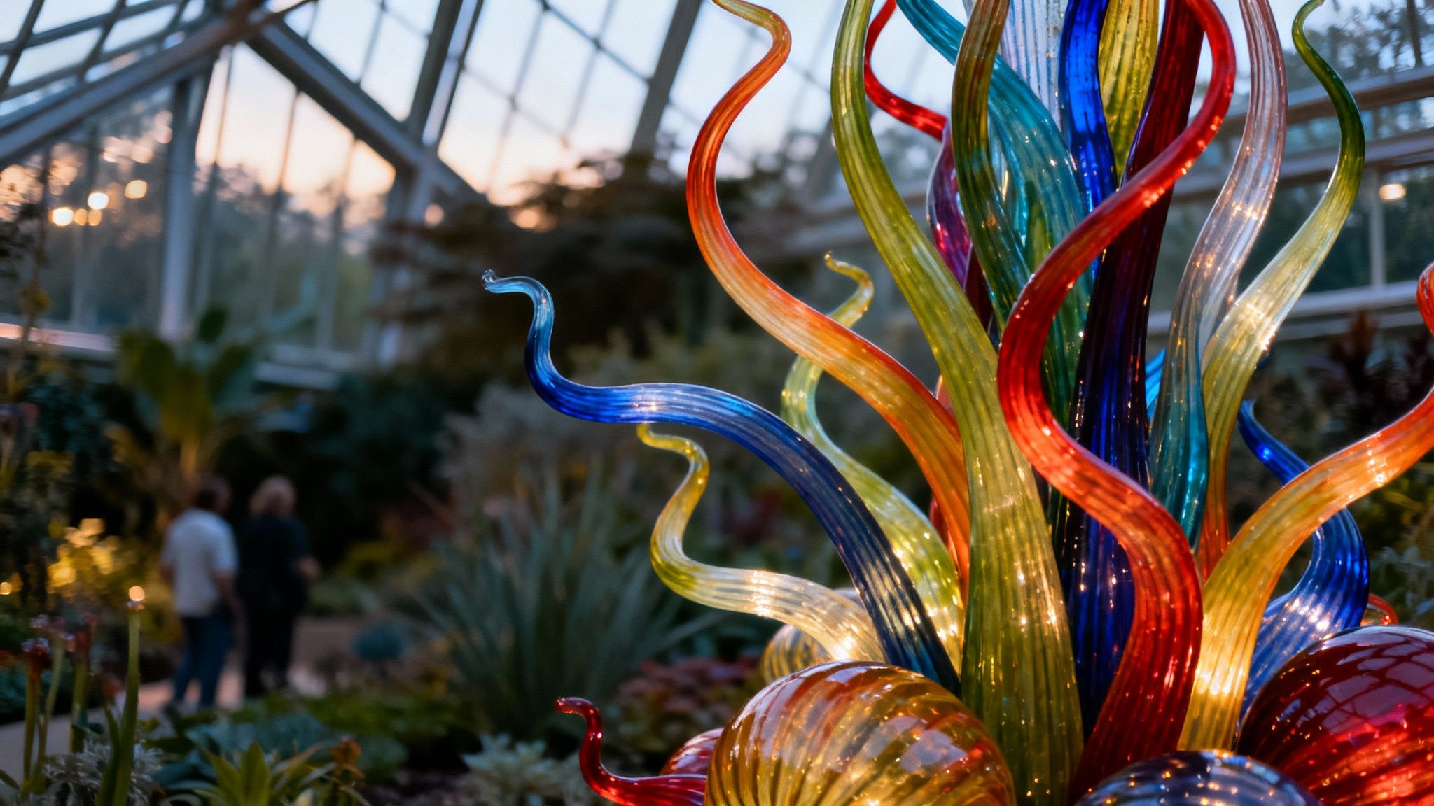 Vibrant Chihuly-style glass sculpture with colorful twisted forms inside a botanical garden greenhouse.