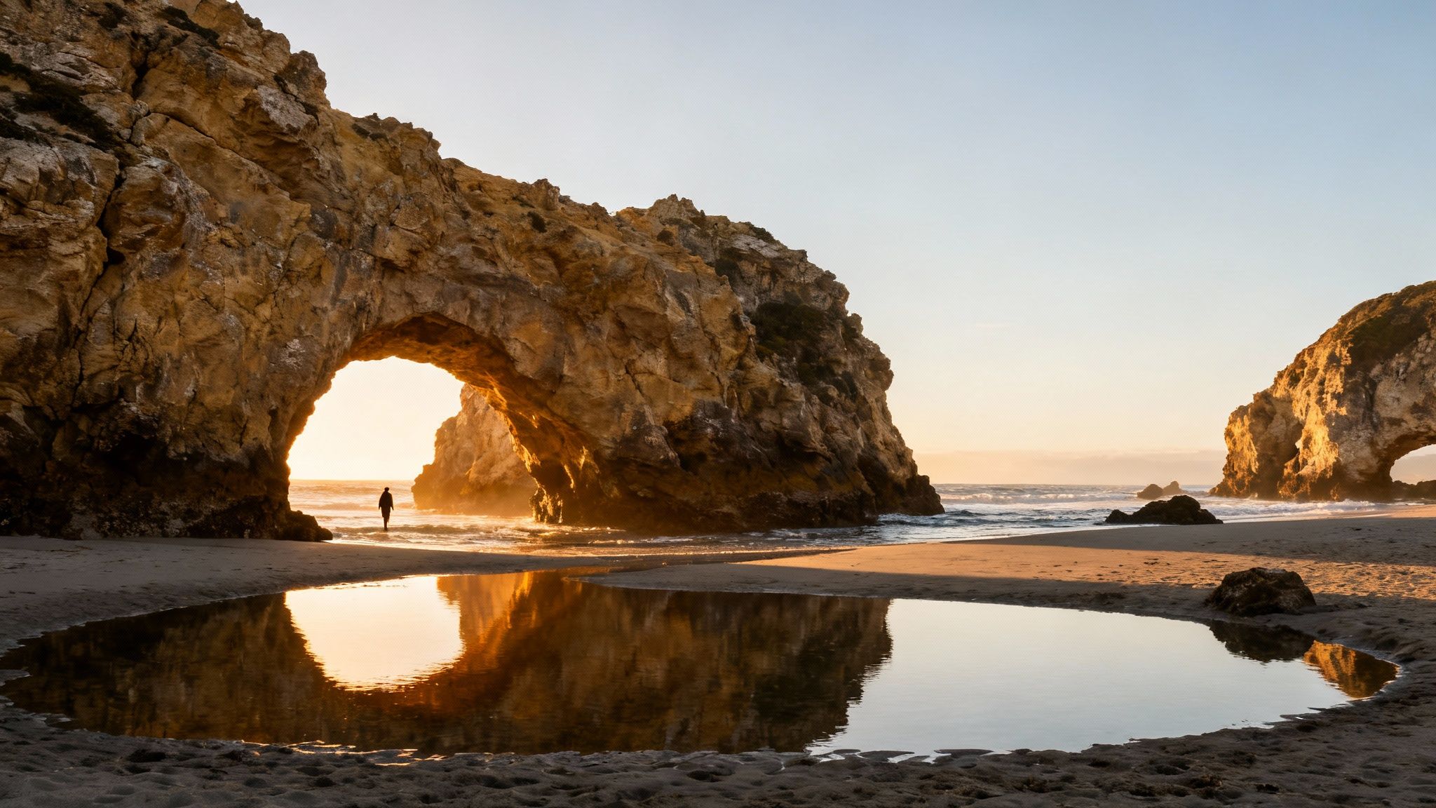 Sunset illuminates a person on a beach through a natural rock arch, reflected in water.